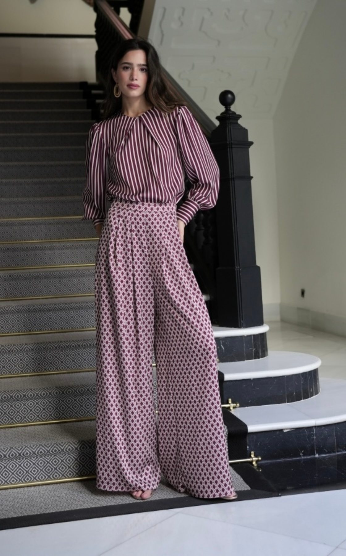 A woman in a maroon and white striped blouse and patterned wide-leg pants standing on a staircase indoors.
