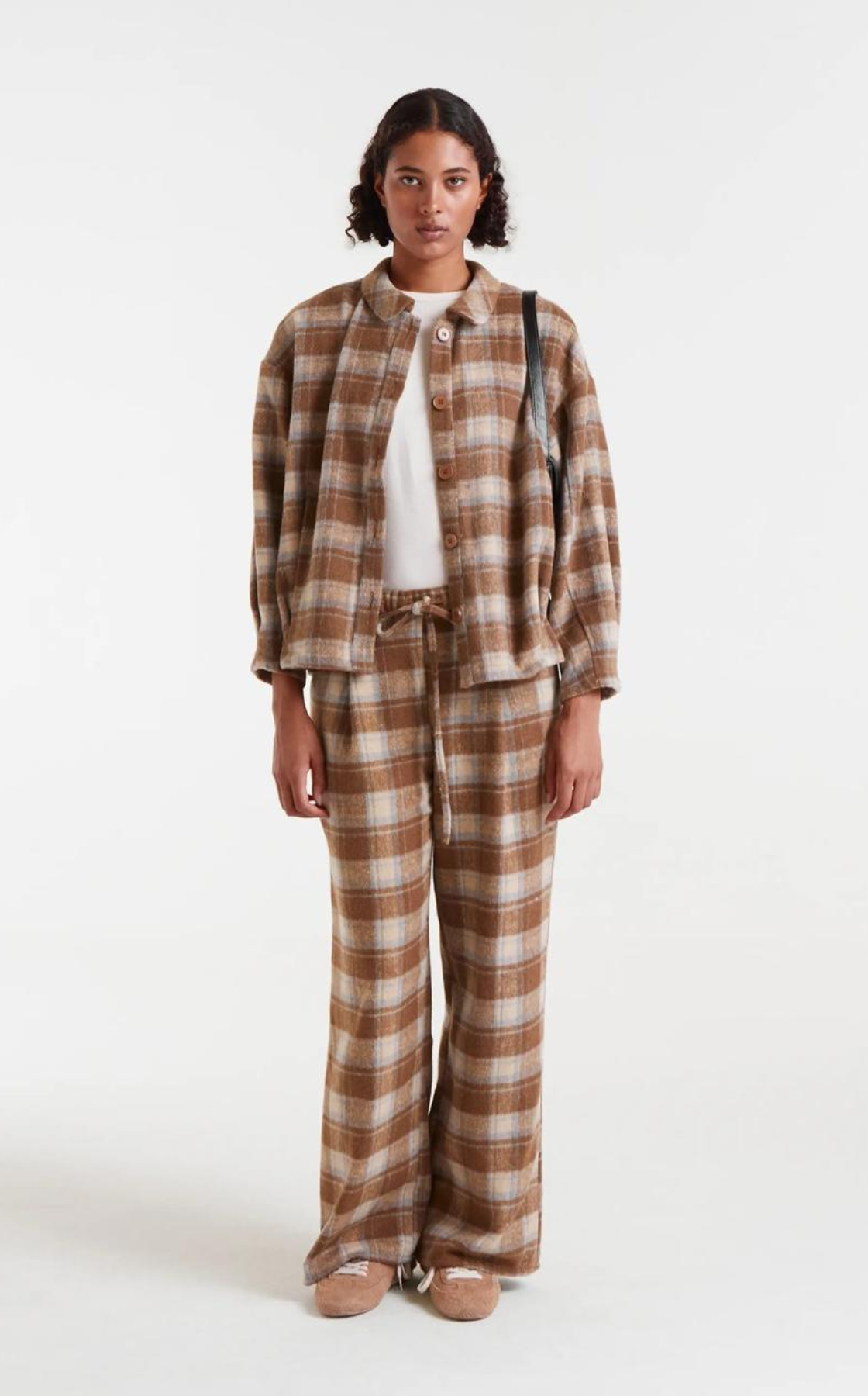 Woman wearing a brown plaid jacket and matching plaid pants, carrying a black shoulder bag, white shirt and beige sneakers, standing against a plain background.