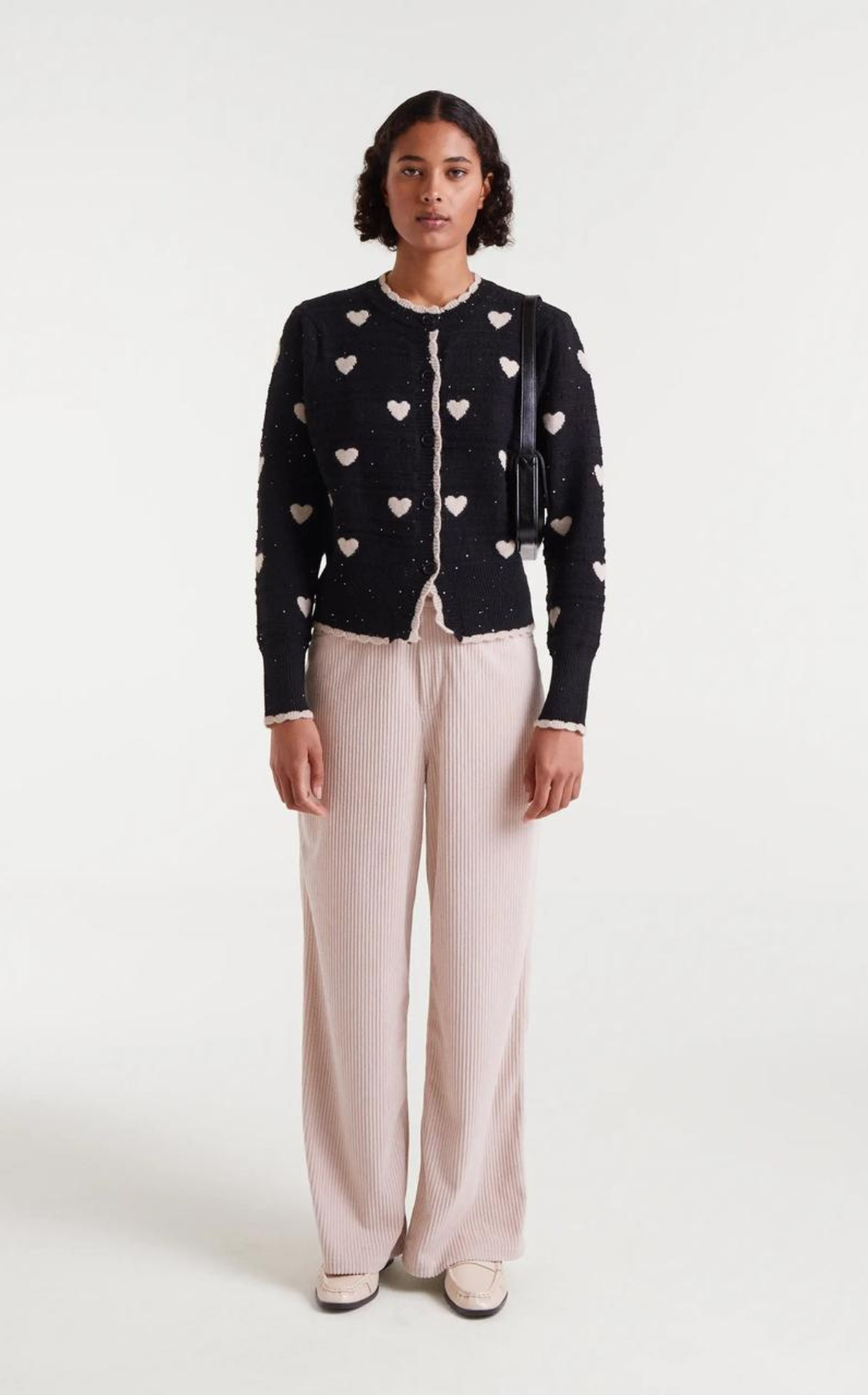 A woman standing in front of a plain white background, wearing a black cardigan with white heart patterns, light-colored wide-leg pants, and beige shoes, holding a black shoulder bag.