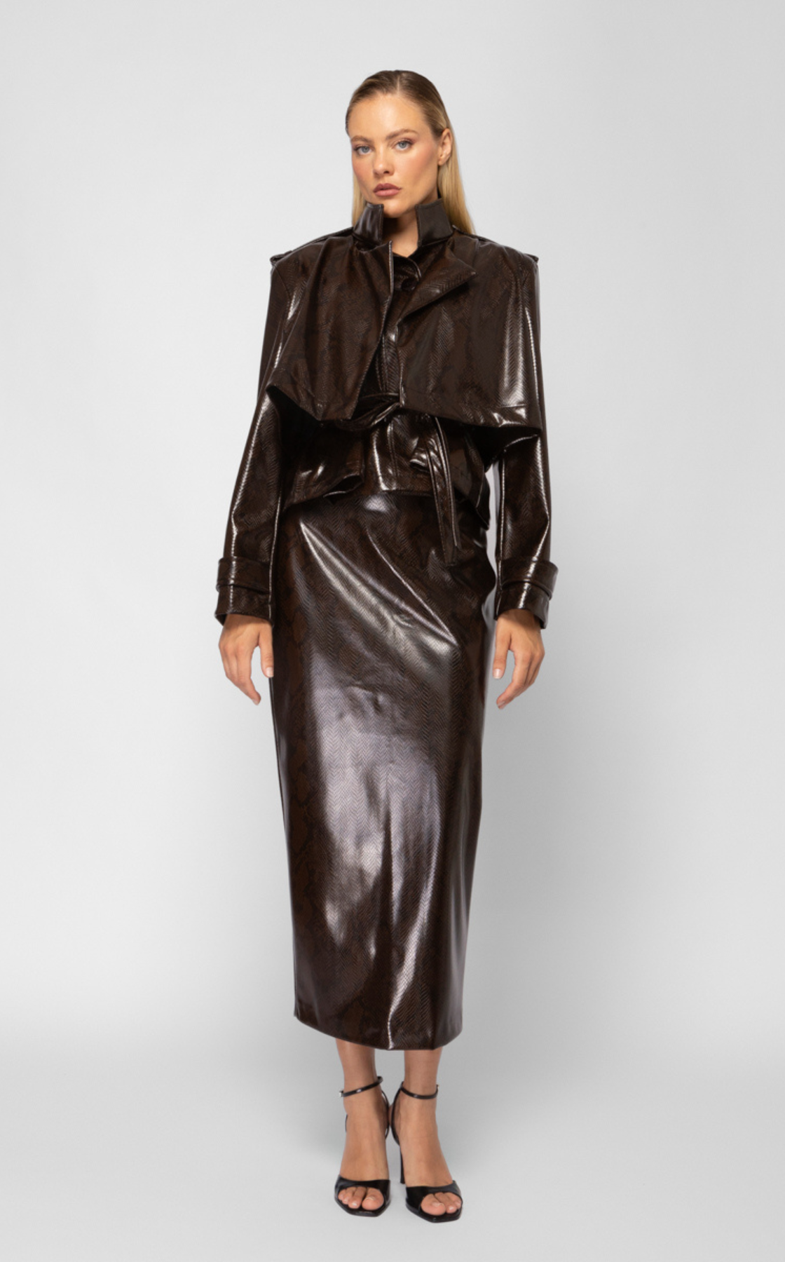 A woman wearing a shiny dark brown leather jacket and matching skirt, paired with black high heels, standing against a plain gray background.