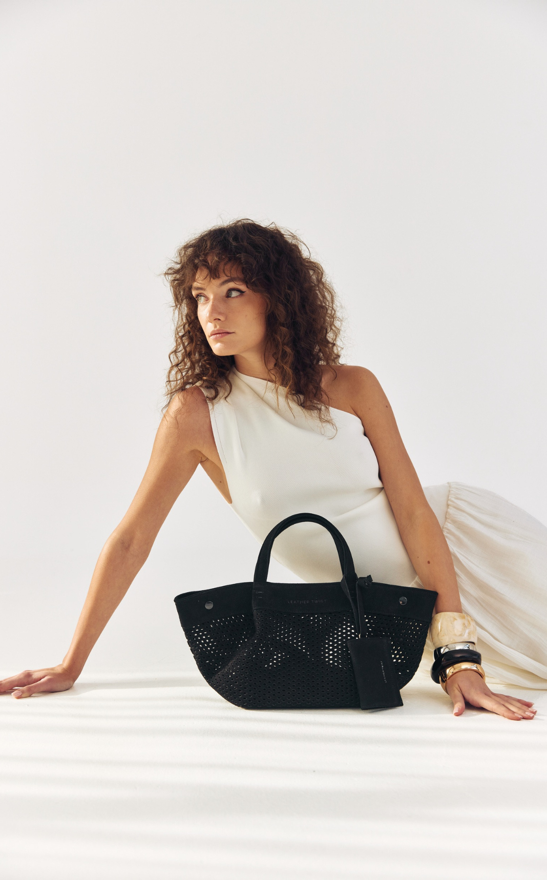 A woman with curly hair in a cream-colored dress sitting on the floor with a black perforated handbag and gold accessories.