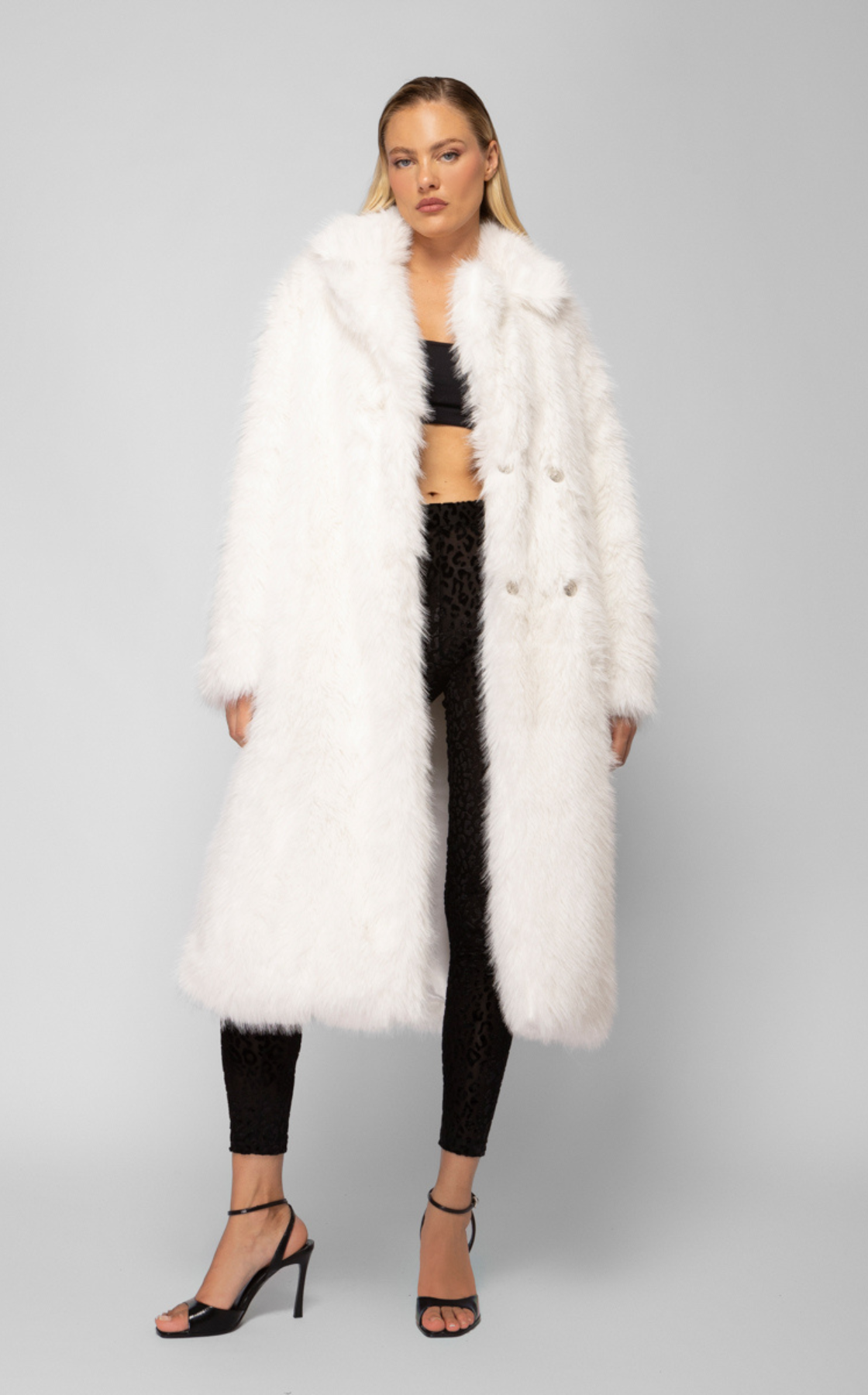 A woman is wearing a long white faux fur coat, black crop top, black leopard print pants, and black high-heeled sandals.