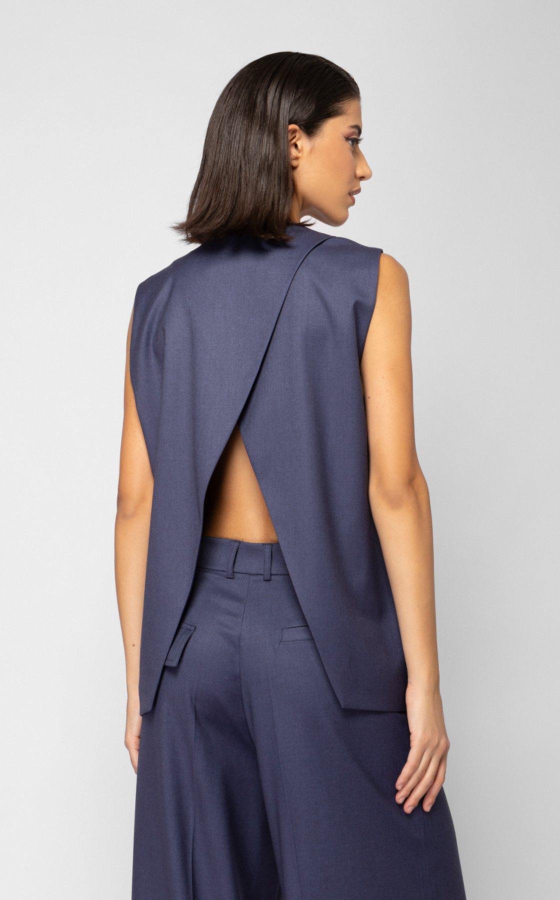 A woman with dark brown, straight hair turned sideways, wearing a blue sleeveless blazer with an open back and matching blue wide-leg pants.