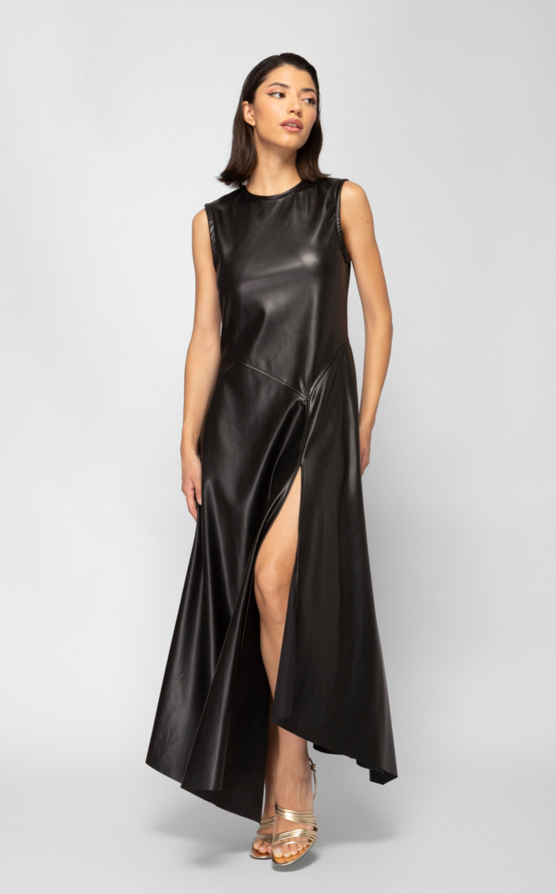A woman wearing a sleeveless, black, floor-length leather dress with a high slit, standing against a plain white background.