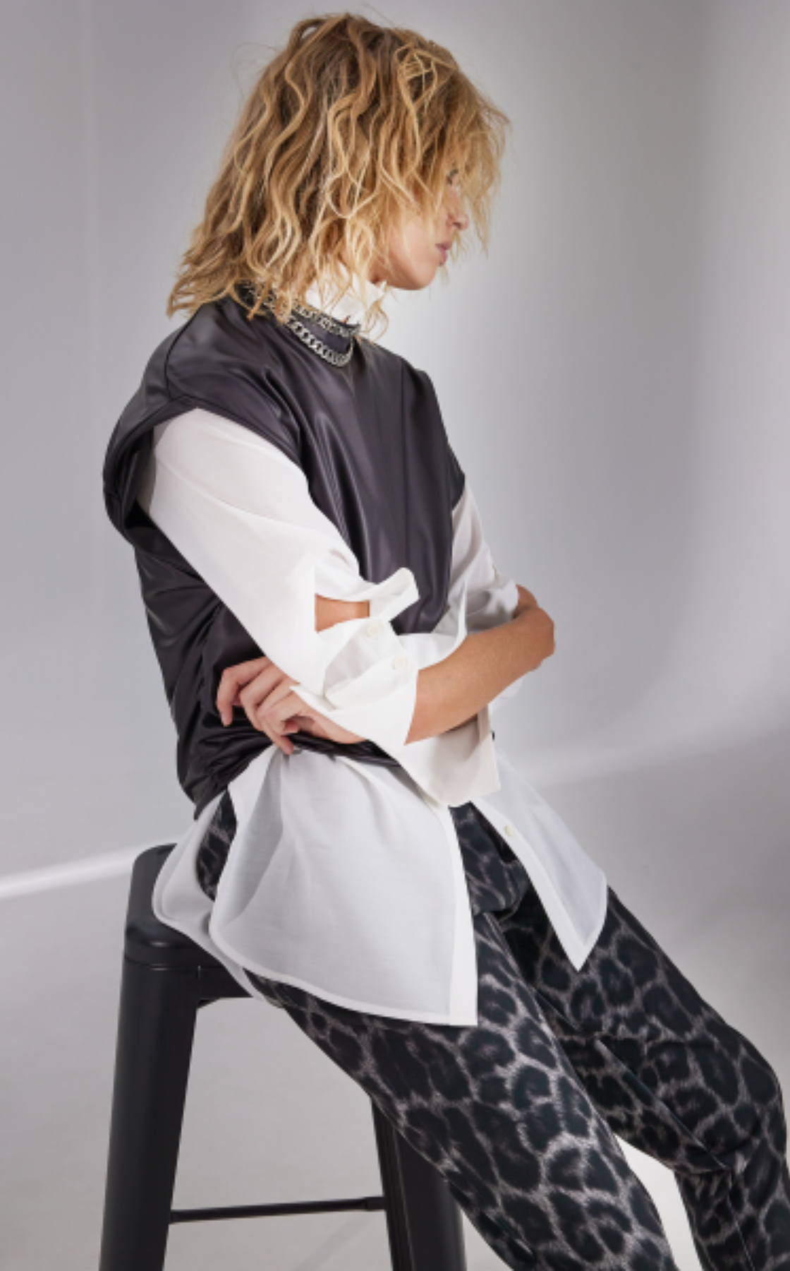 A woman with curly blonde hair sitting on a black stool, wearing a white shirt layered under a black sleeveless vest and leopard print pants.
