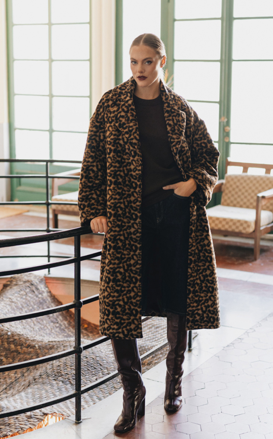 A woman in a long leopard print coat, black top, dark jeans, and high-heeled boots standing indoors near railing with large windows in the background.