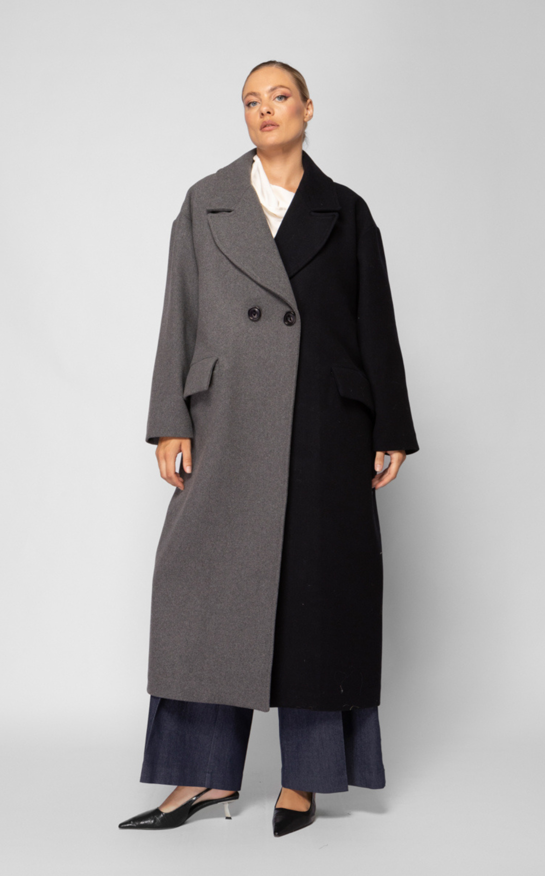 A woman wearing a long, two-tone coat with one side gray and the other black, standing against a plain light gray background.