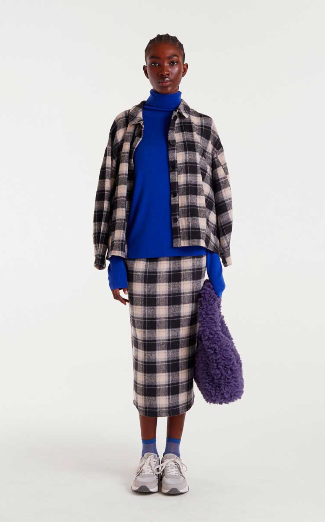 A woman wearing a blue turtleneck, plaid skirt, and layered plaid shirt, holding a purple fuzzy bag, with white sneakers and gray socks, standing against a plain white background.