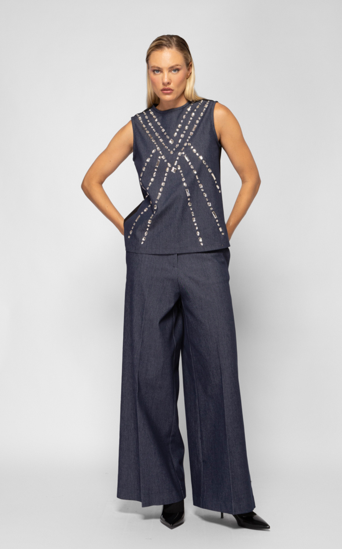 A woman with blond hair wearing a sleeveless dark denim top with silver embellishments and matching wide-leg denim pants, standing against a plain light background.