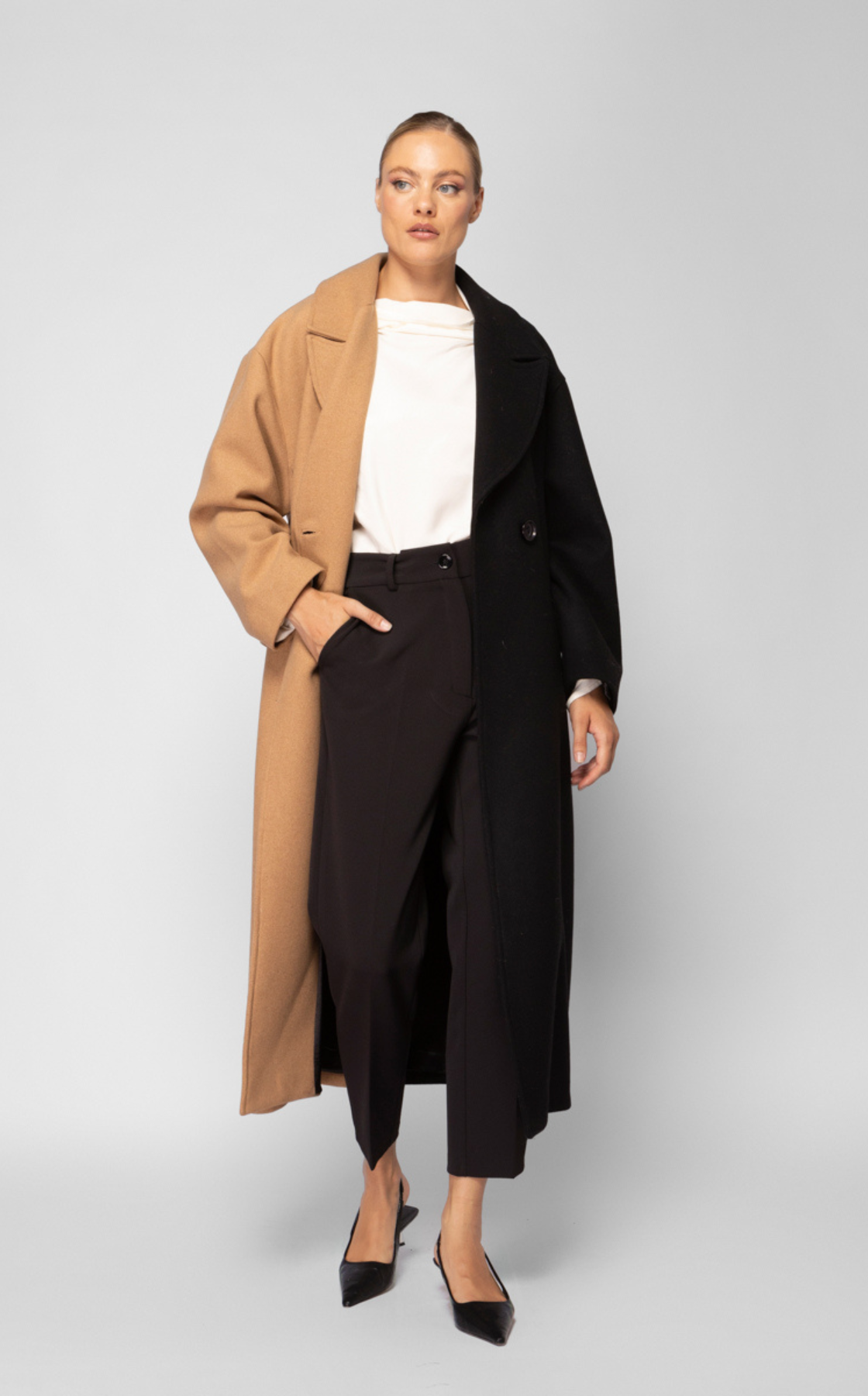 A woman wearing a black and tan color-block coat, black high-waisted trousers, a white blouse, and black pointed-toe heels.