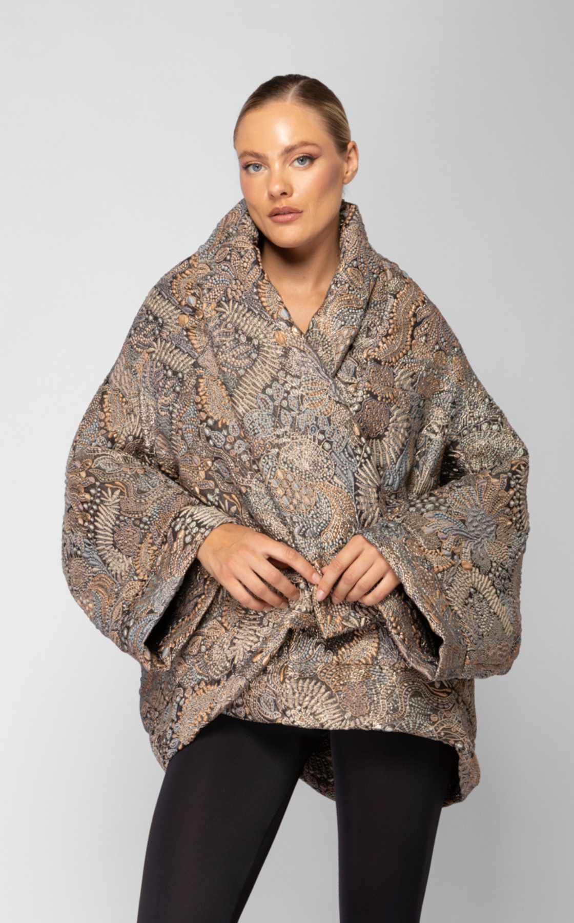 A woman with blonde hair styled back, wearing a patterned, textured coat with earth-tone colors and black pants, standing against a plain white background.