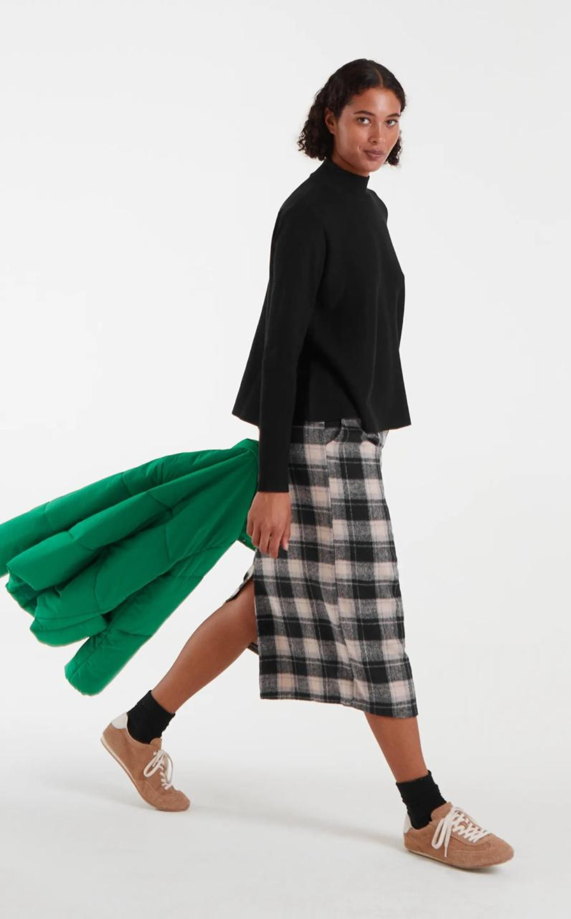 Woman walking, wearing a black top, plaid skirt, beige sneakers, and black socks, with a green jacket tucked under her arm.