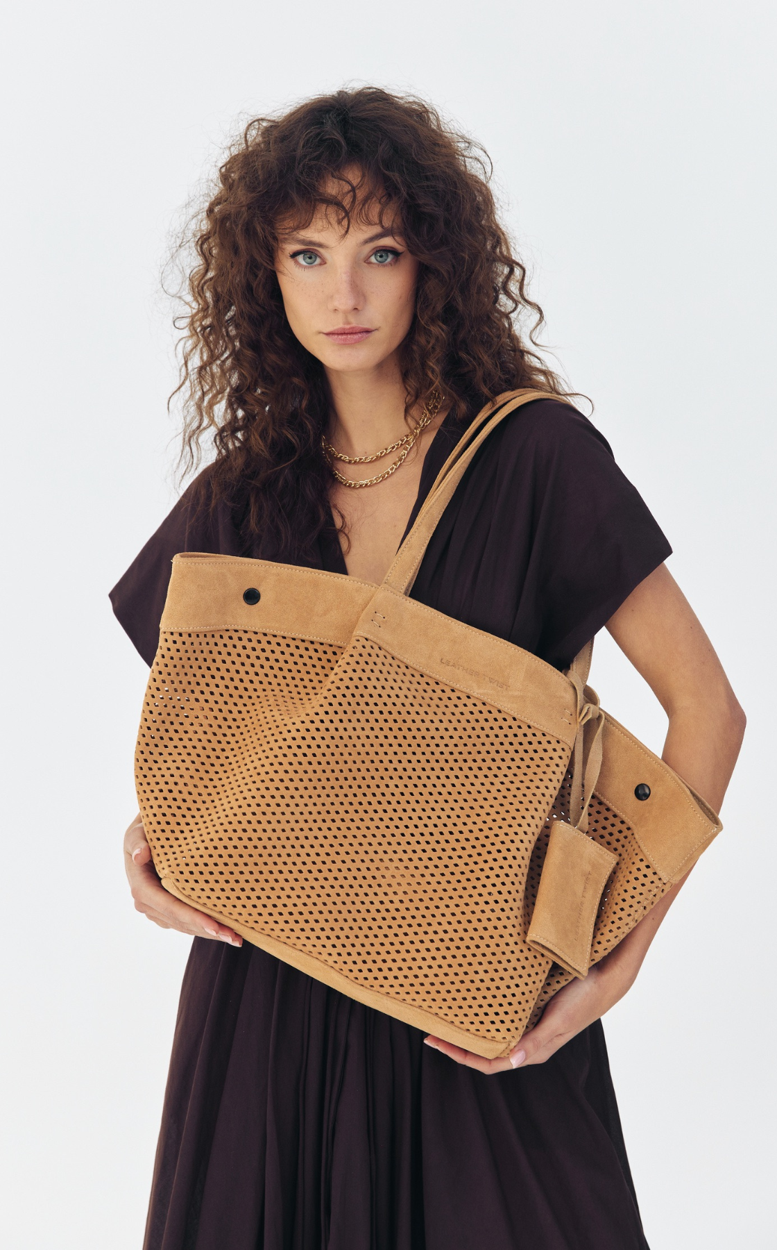 Woman with curly brown hair wearing a black dress and multiple gold necklaces, holding a large tan mesh tote bag with leather accents against a plain white background.