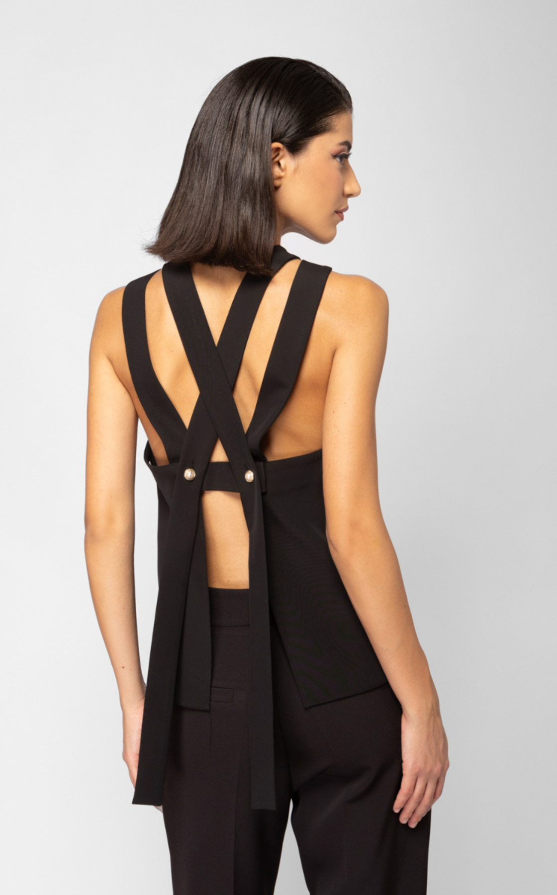 A woman with shoulder-length dark hair wearing a black backless top with wide straps and black pants, standing against a plain gray background.