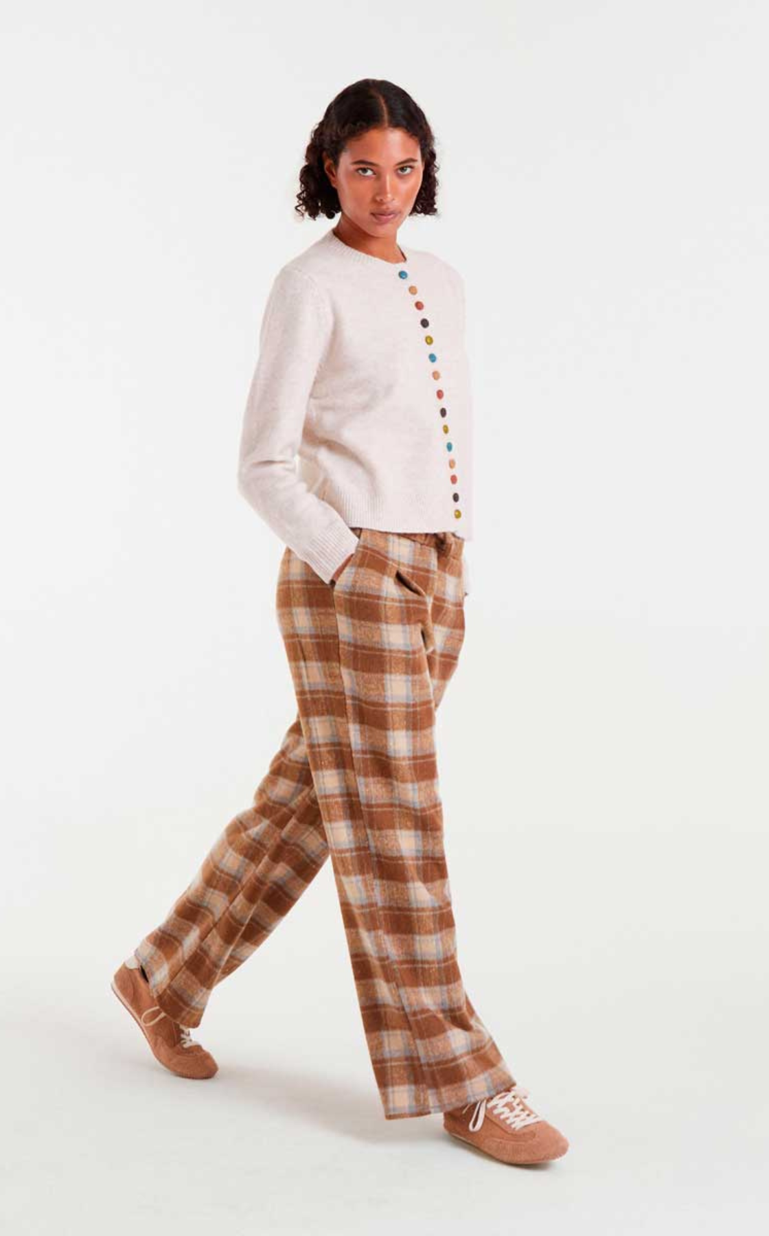 Woman wearing a beige cardigan with multicolored buttons and plaid wide-leg pants, standing with her hands in her pockets against a plain white background.