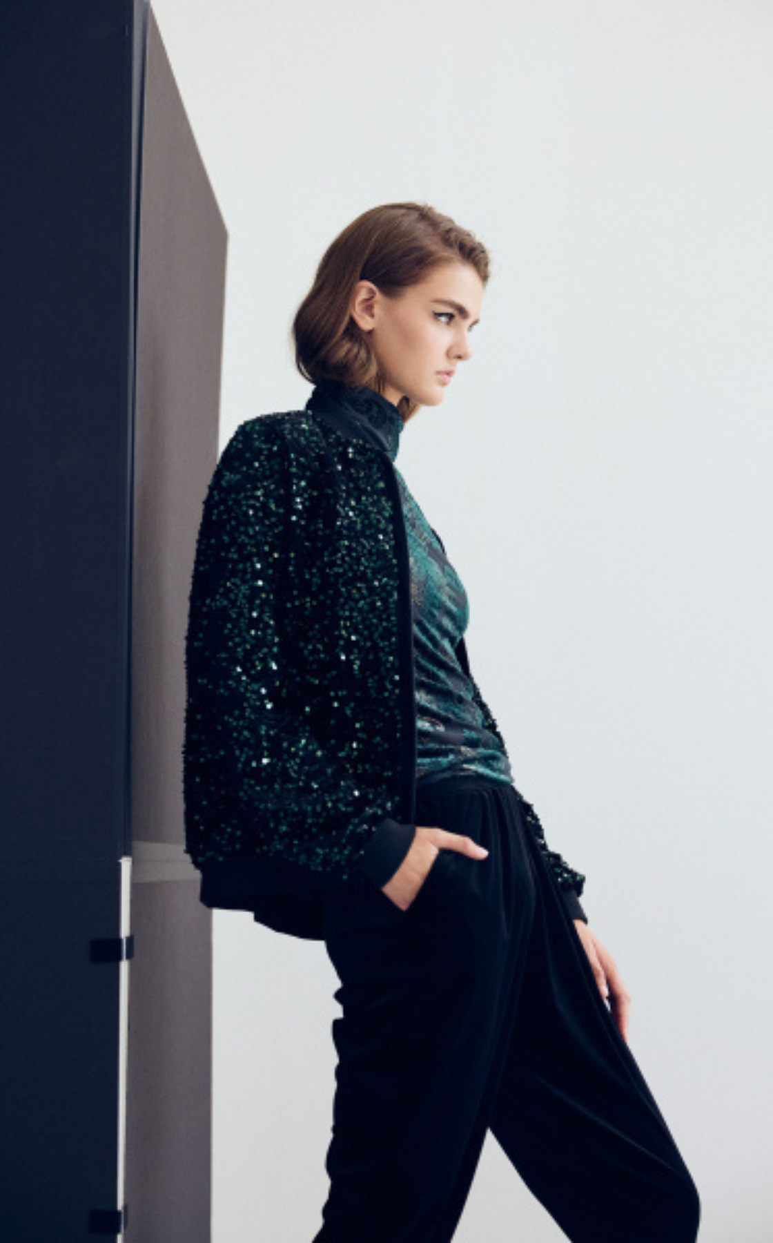 Fashion model in sequined jacket and black pants poses against gray wall.