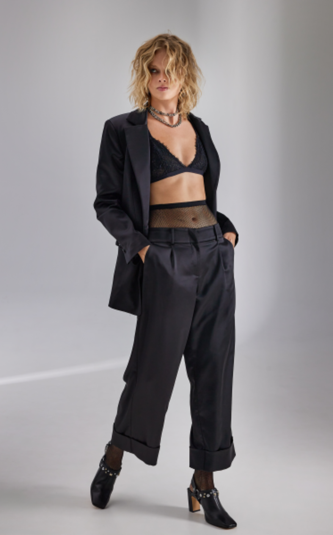 Woman with curly blonde hair wearing a black blazer, black lace top, fishnet midriff, black trousers, and black heeled boots with gold studs, against a plain gray background.