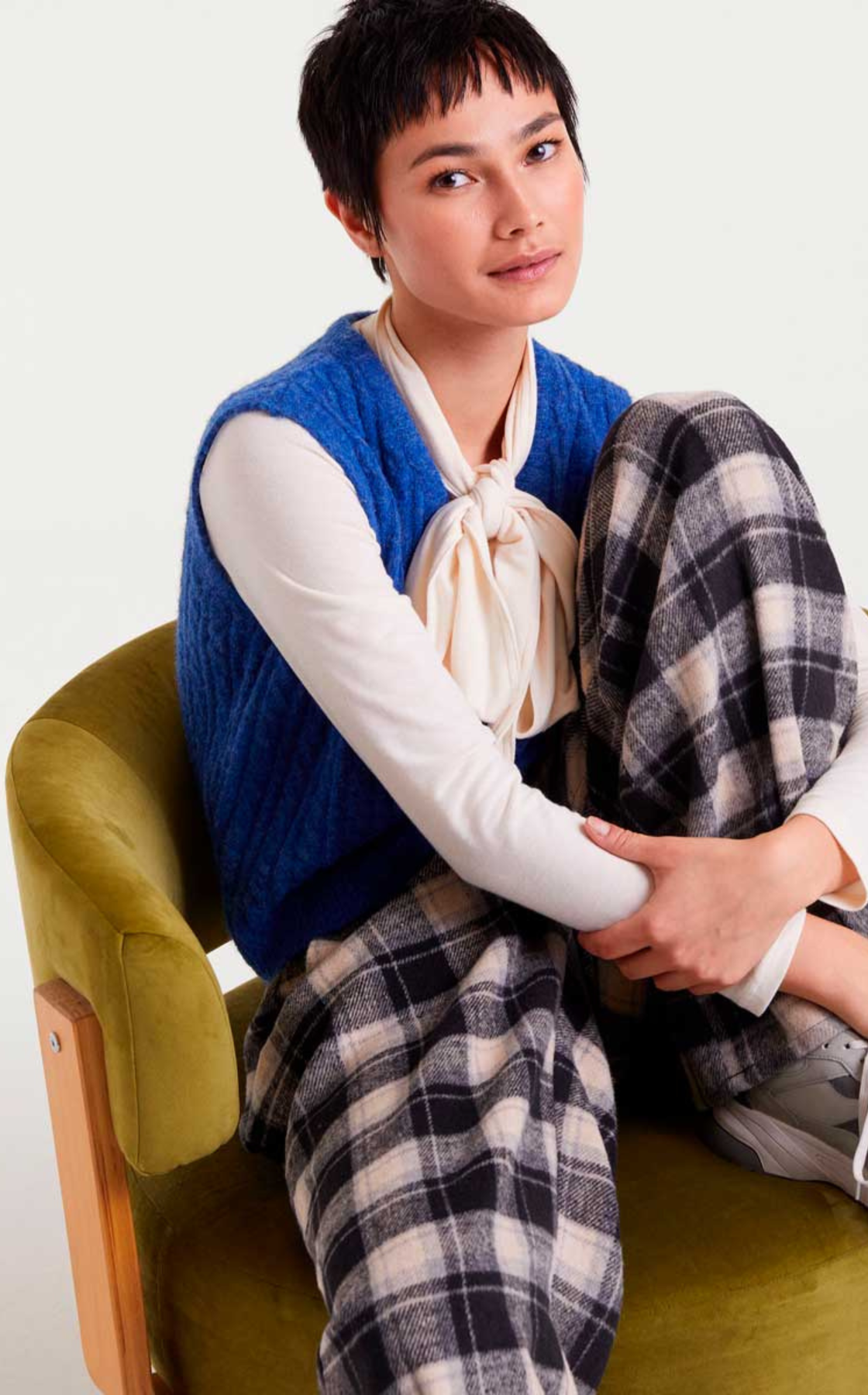 A woman with short dark hair sitting on an olive green chair, wearing a blue vest over a white blouse with a bow, and plaid pants, in a studio setting.