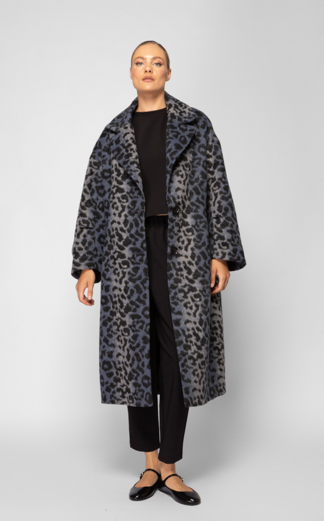 A woman wearing a long, leopard-print coat with a black shirt, black pants, and black shoes stands against a plain background.