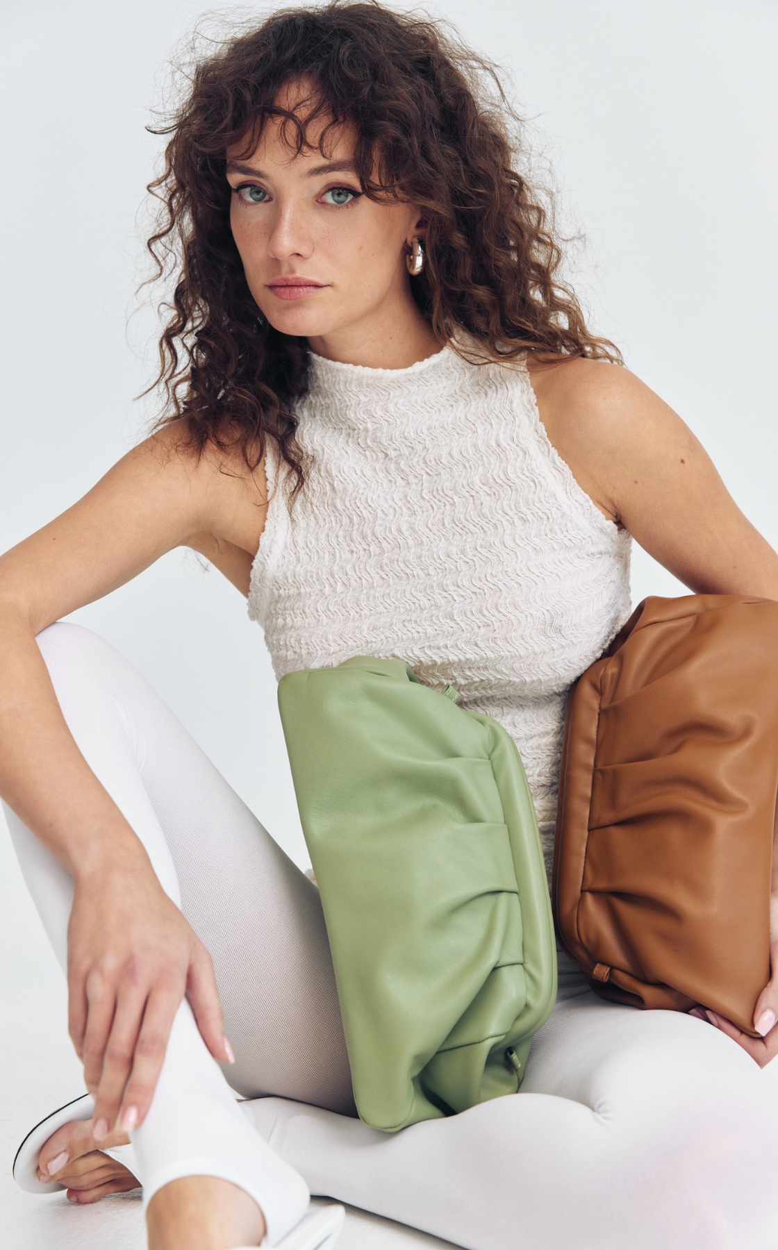 A woman with curly, shoulder-length hair and blue eyes, wearing a sleeveless white textured top, beige pants, and white shoes. She is sitting on a white surface with two handbags, one green and one brown, placed on her lap.