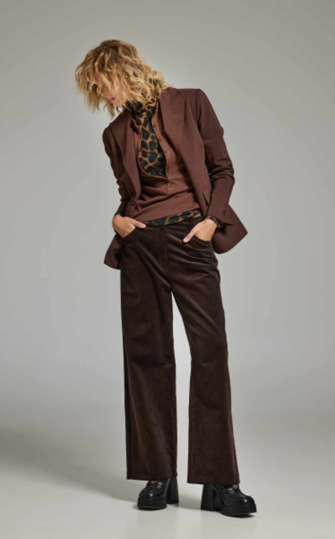 A woman with curly blonde hair wearing a brown blazer, leopard print turtleneck, brown corduroy pants, and black platform boots, standing against a plain background.