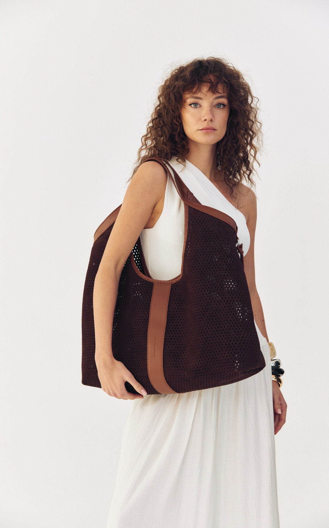 Woman with curly hair wearing a white top and white skirt, carrying a large brown mesh tote bag with brown straps.