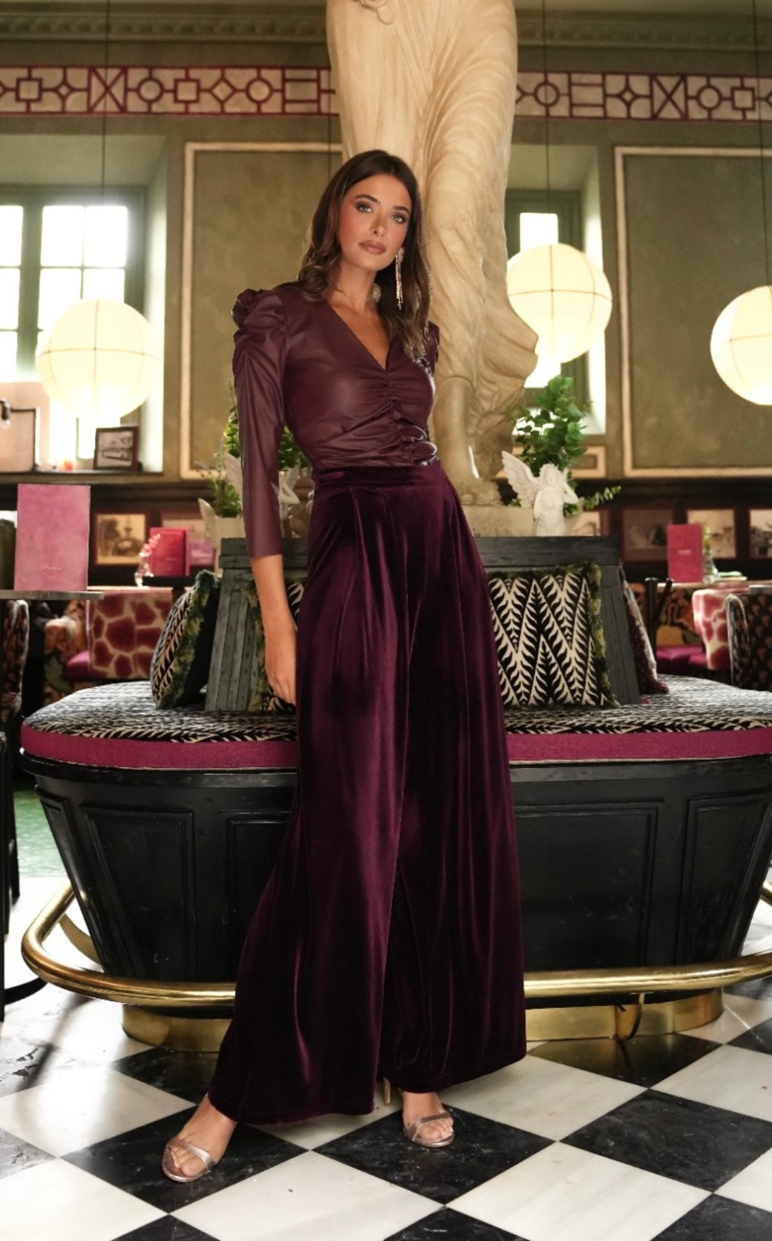 A woman in a burgundy satin top and velvet wide-leg pants standing inside a decorated room.