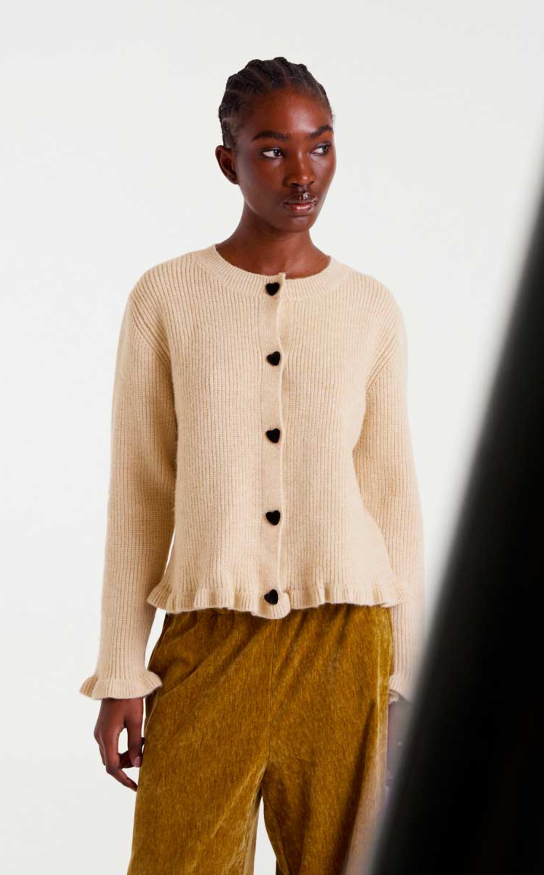 A woman with braided hair wearing a cream-colored knit cardigan with black heart-shaped buttons and ruffle trim at the hem, paired with brown textured pants, standing against a plain white background.