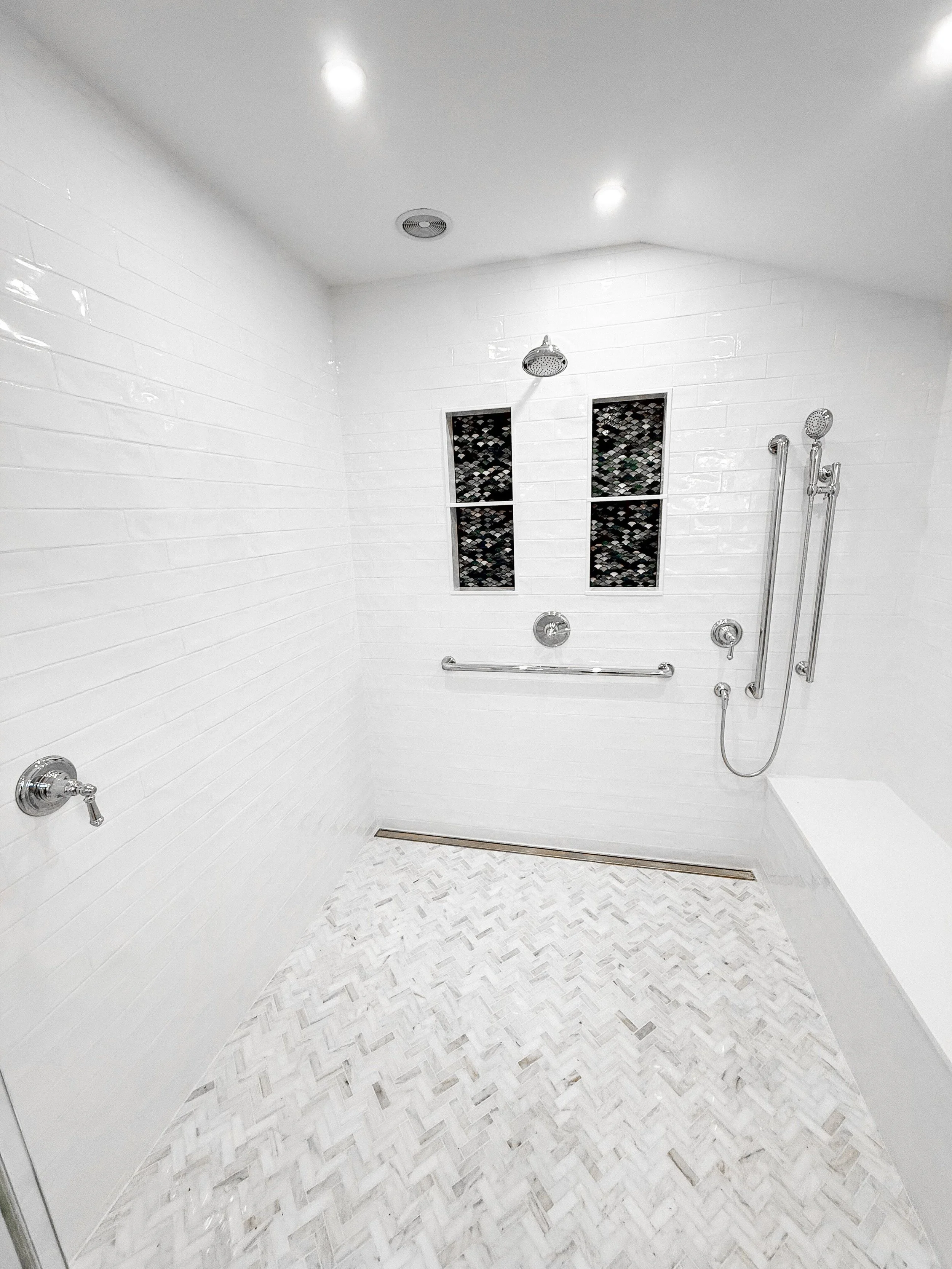 large white shower with patterned tile floor and white tile walls and silver shower heads and accessories plus bench