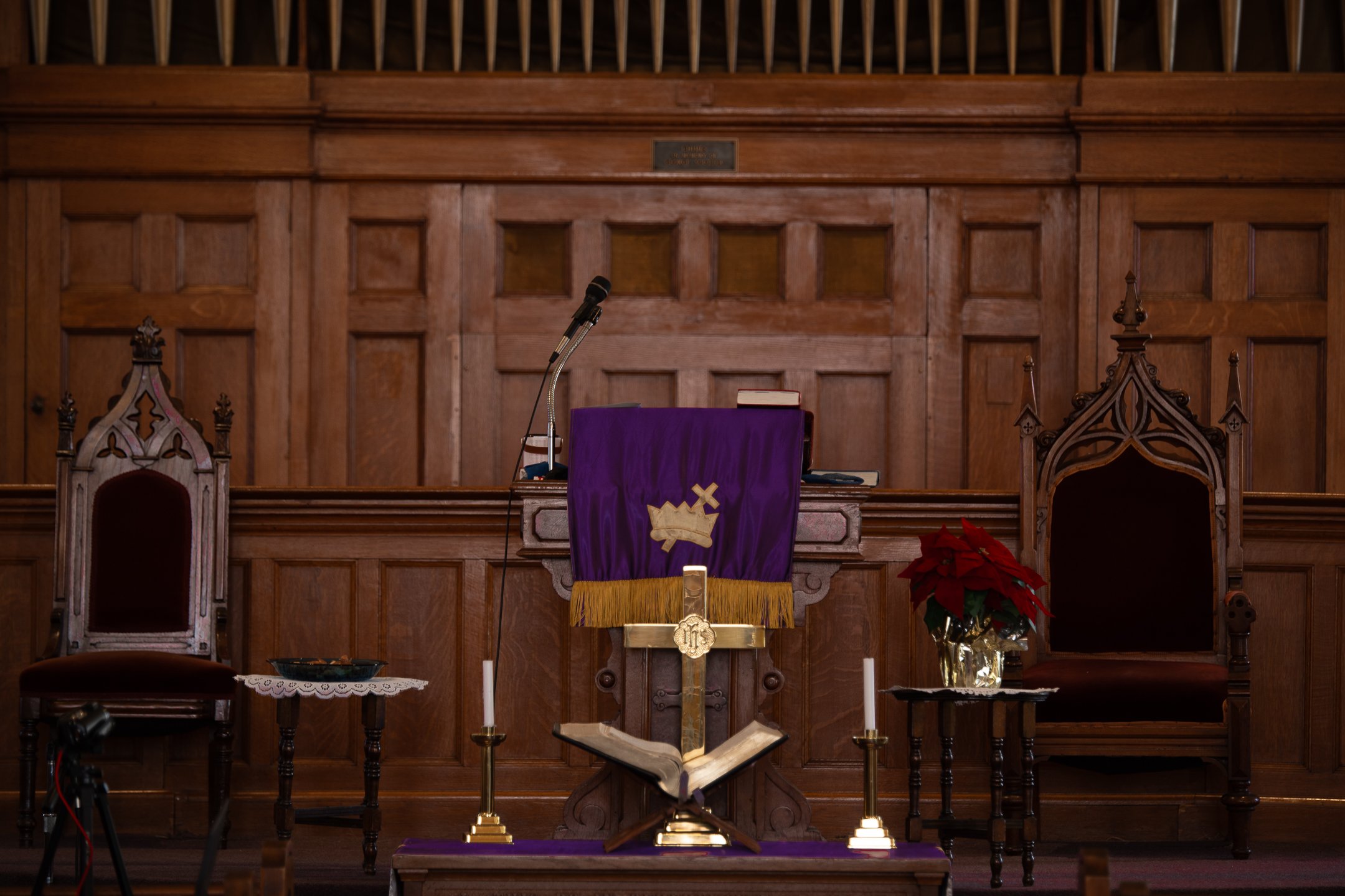 Church pulpit with a purple and gold cloth, microphone, and open Bible, flanked by ornate wooden chairs and poinsettia flowers.