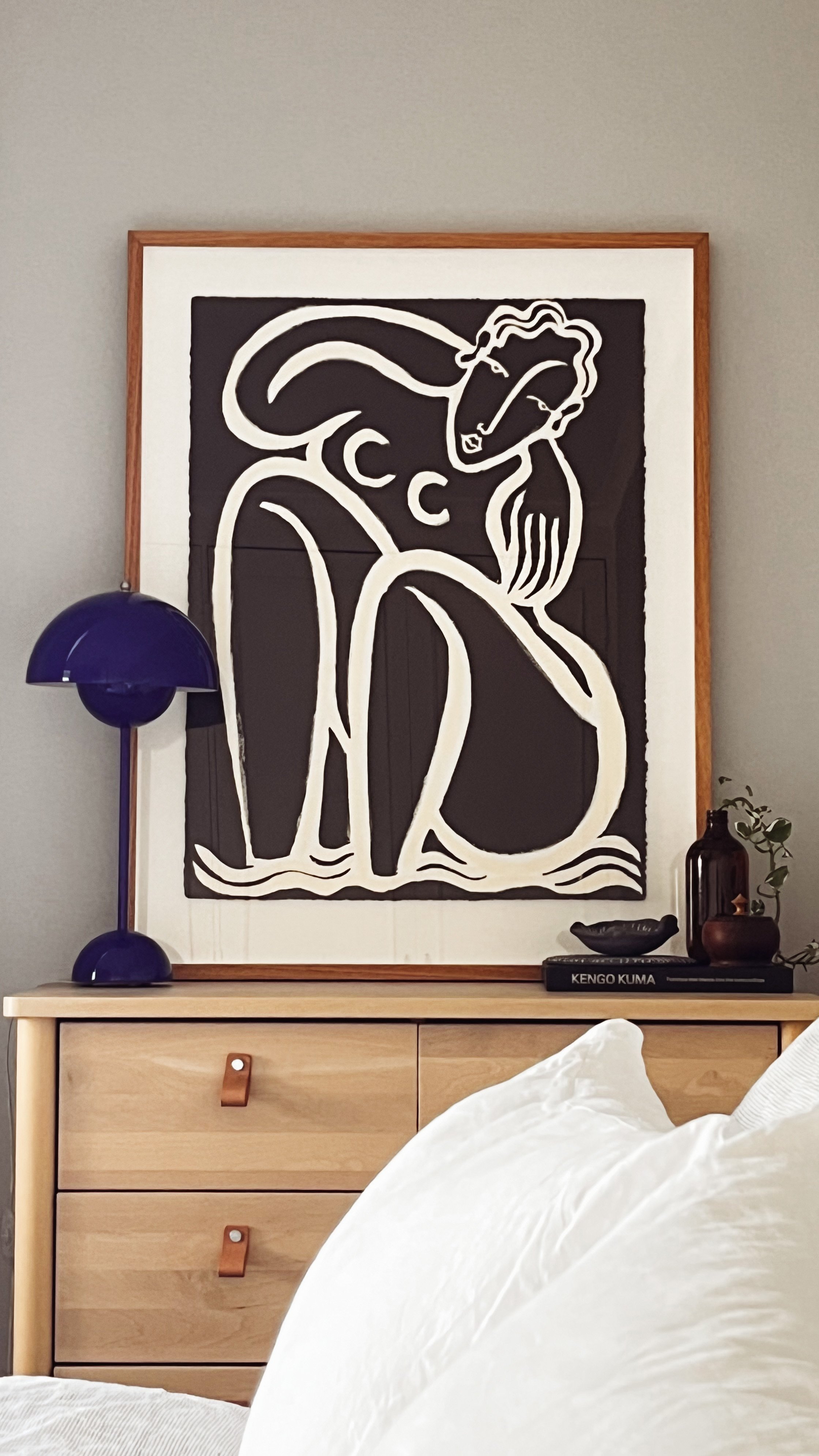 A framed black and white abstract line drawing of a woman sitting, with a blue table lamp to the left, on a light wood dresser with two drawers, and decorative items including a brown glass vase, a small black dish, and a book titled "KENGO KUMA" on top.