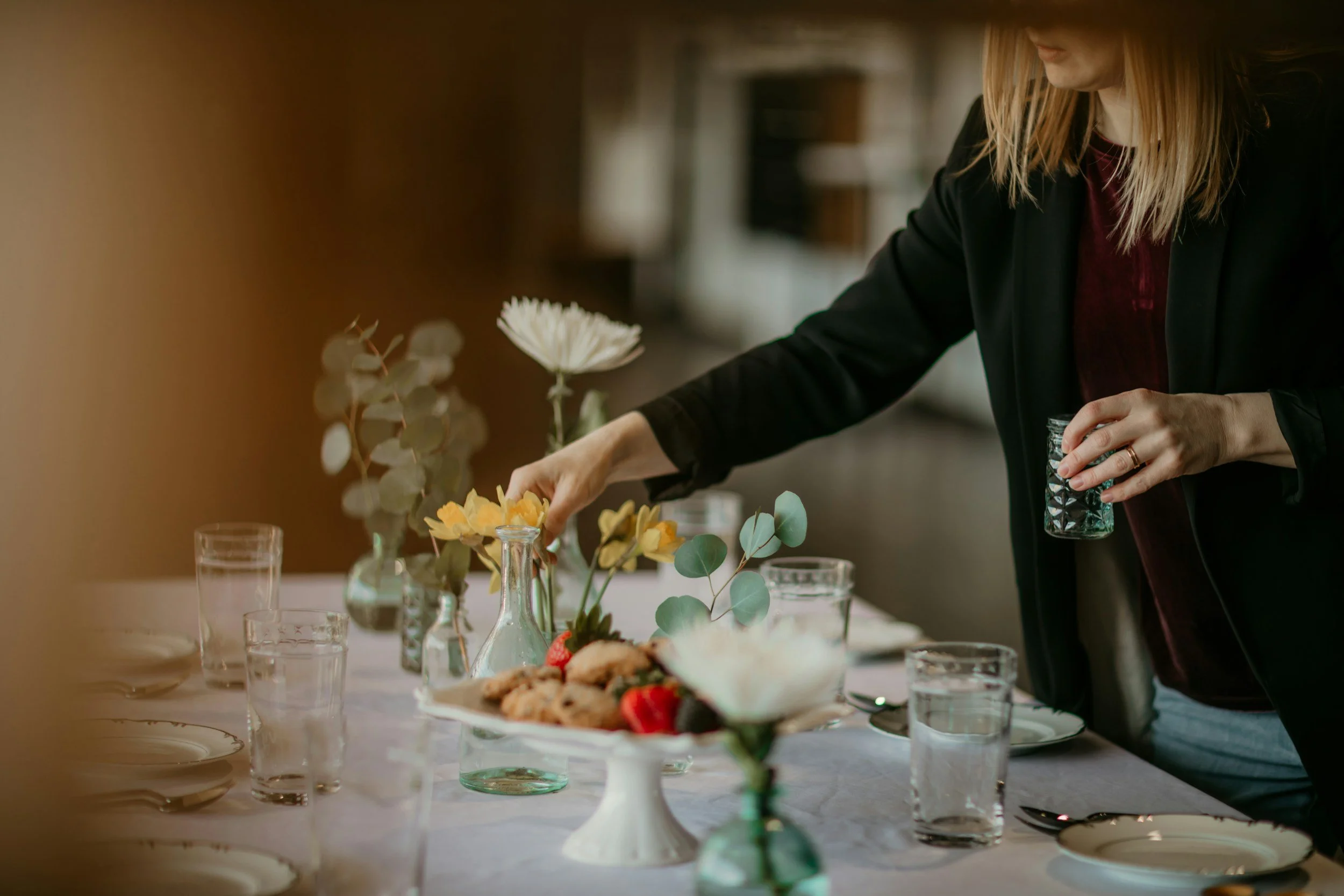 A woman arranging flowers on a table set for a meal, with glasses, plates, and a cake stand with desserts.