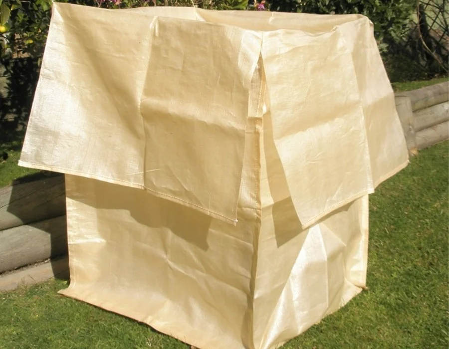 A beige lightweight fabric or paper tent set up outdoors on grass, with a wooden fence and shrubbery in the background.