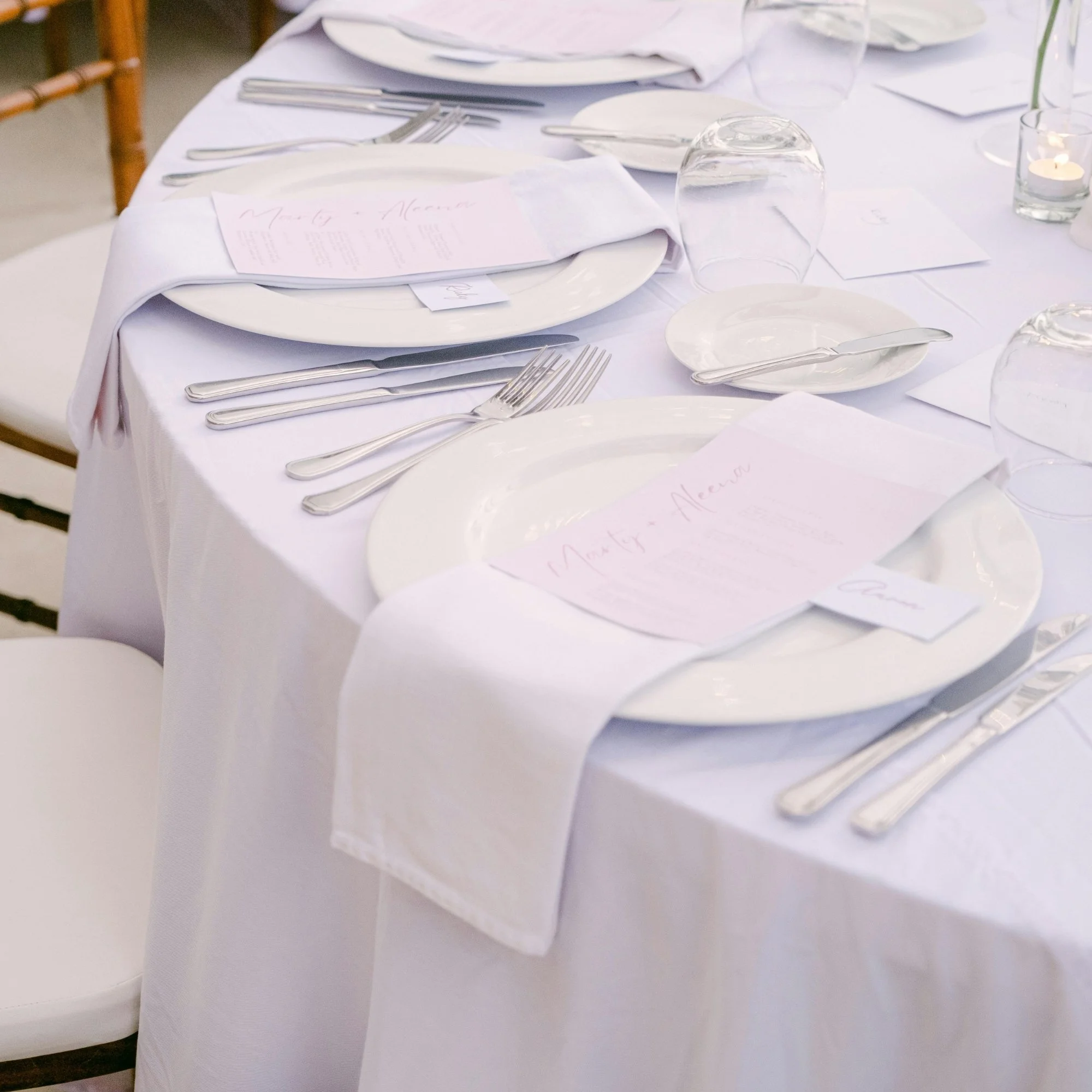 A neatly set dining table with white tablecloth, white plates, silverware, pink place cards, upside-down glasses, and small candles, prepared for an event or celebration.