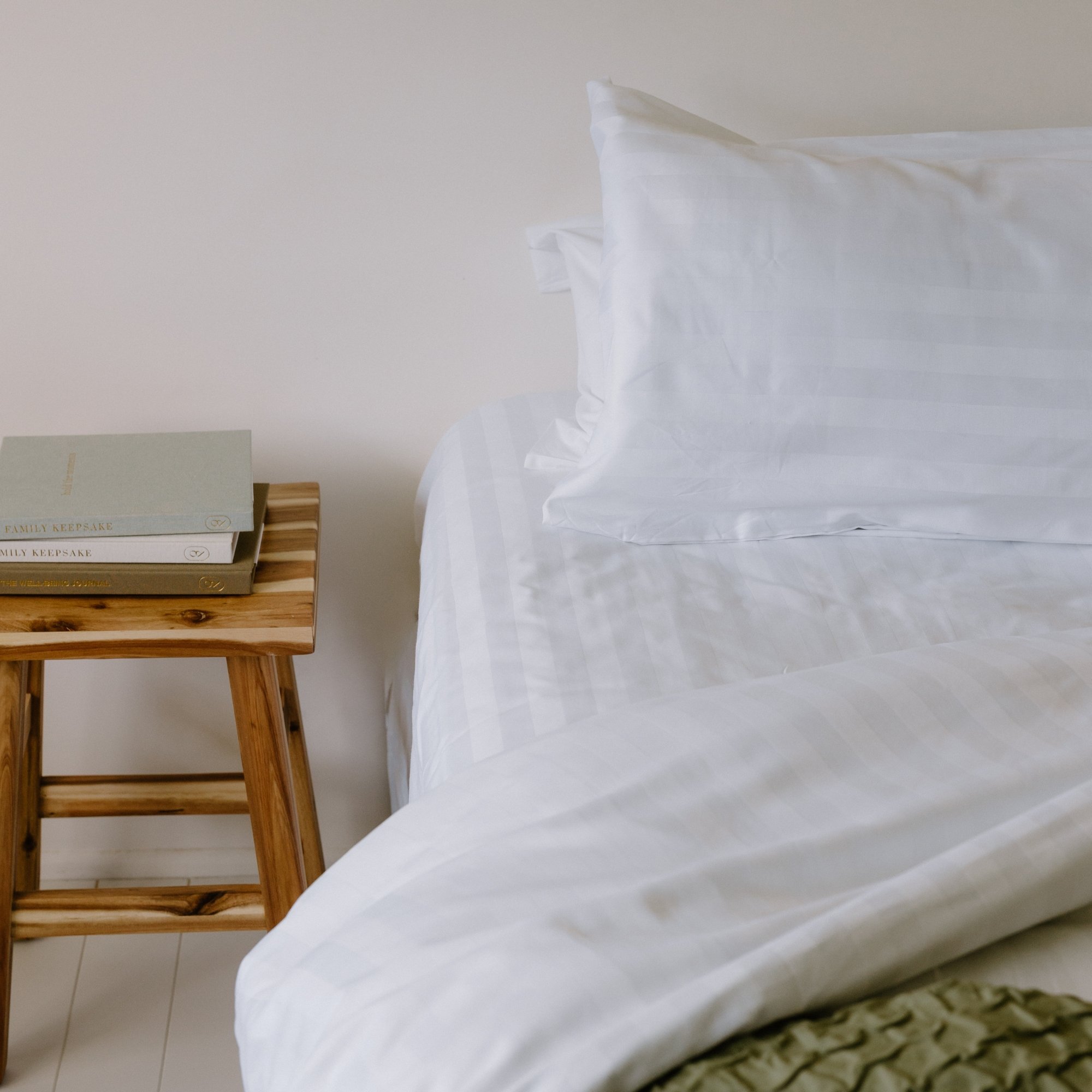 A neatly made bed with white striped sheets and pillows, next to a wooden nightstand with stacked books on top.