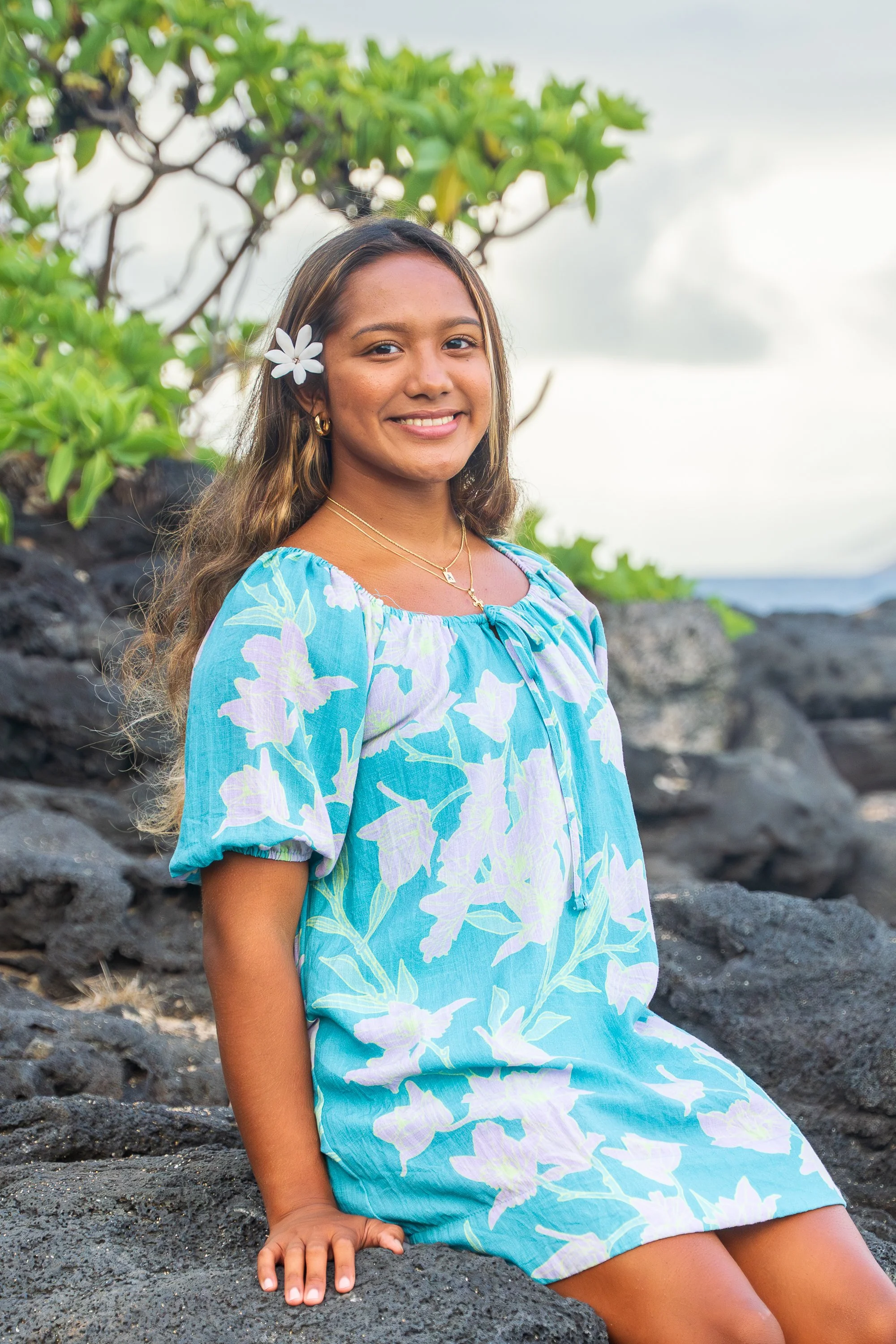 A young woman sits on black volcanic rocks on the beach, smiling, with greenery and cloudy sky in the background. She wears a blue and white floral dress, gold jewelry, and a white flower in her hair.