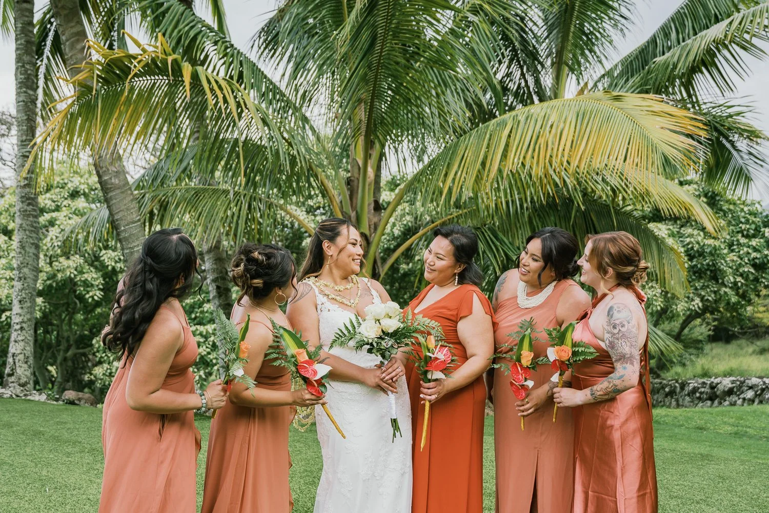 Group of women in wedding attire standing outdoors under palm trees, smiling and holding bouquets of flowers.