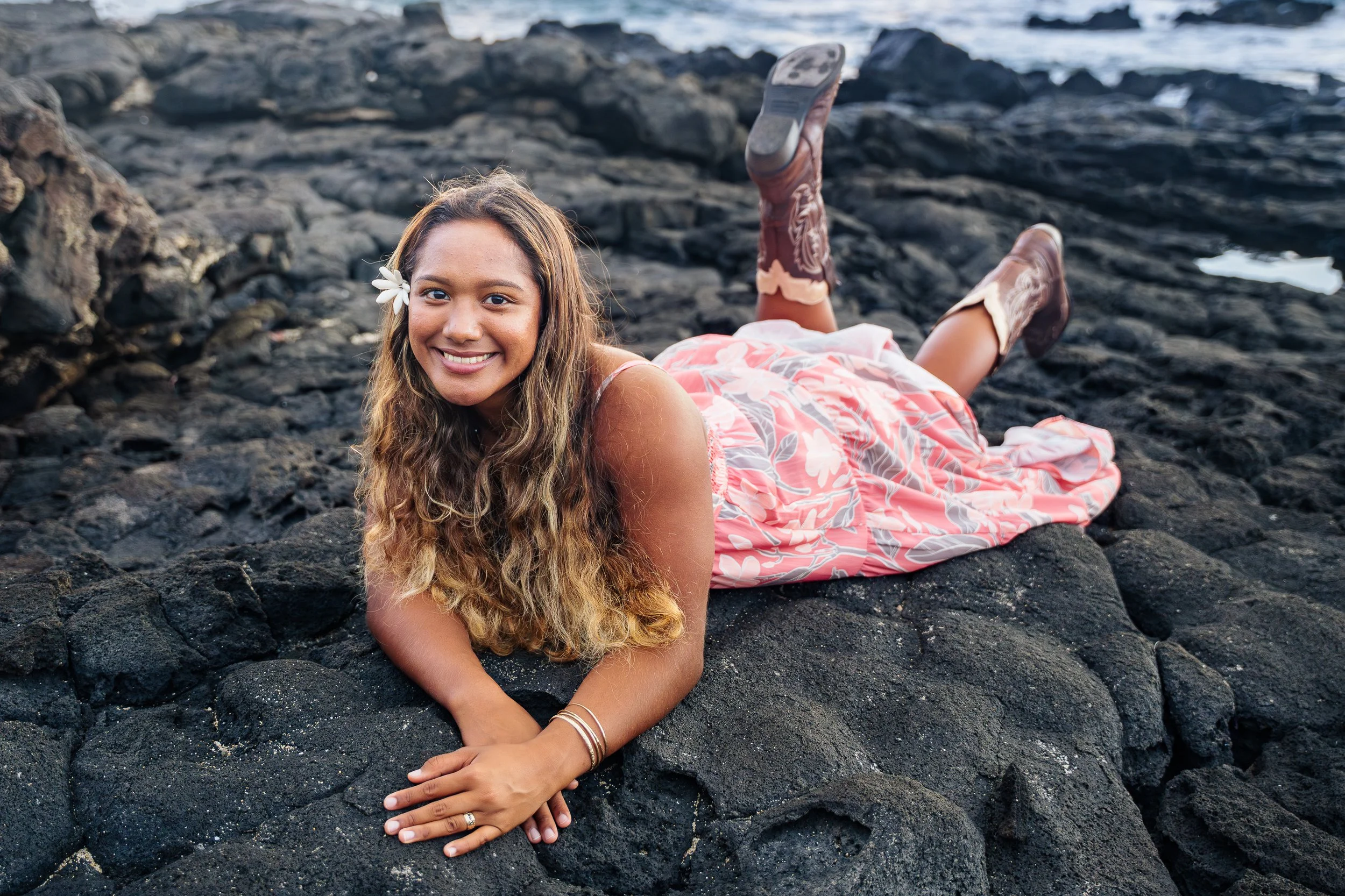 A woman with long wavy hair, wearing a pink floral dress and cowboy boots, lying on black volcanic rocks near the ocean, smiling at the camera with a white flower in her hair.