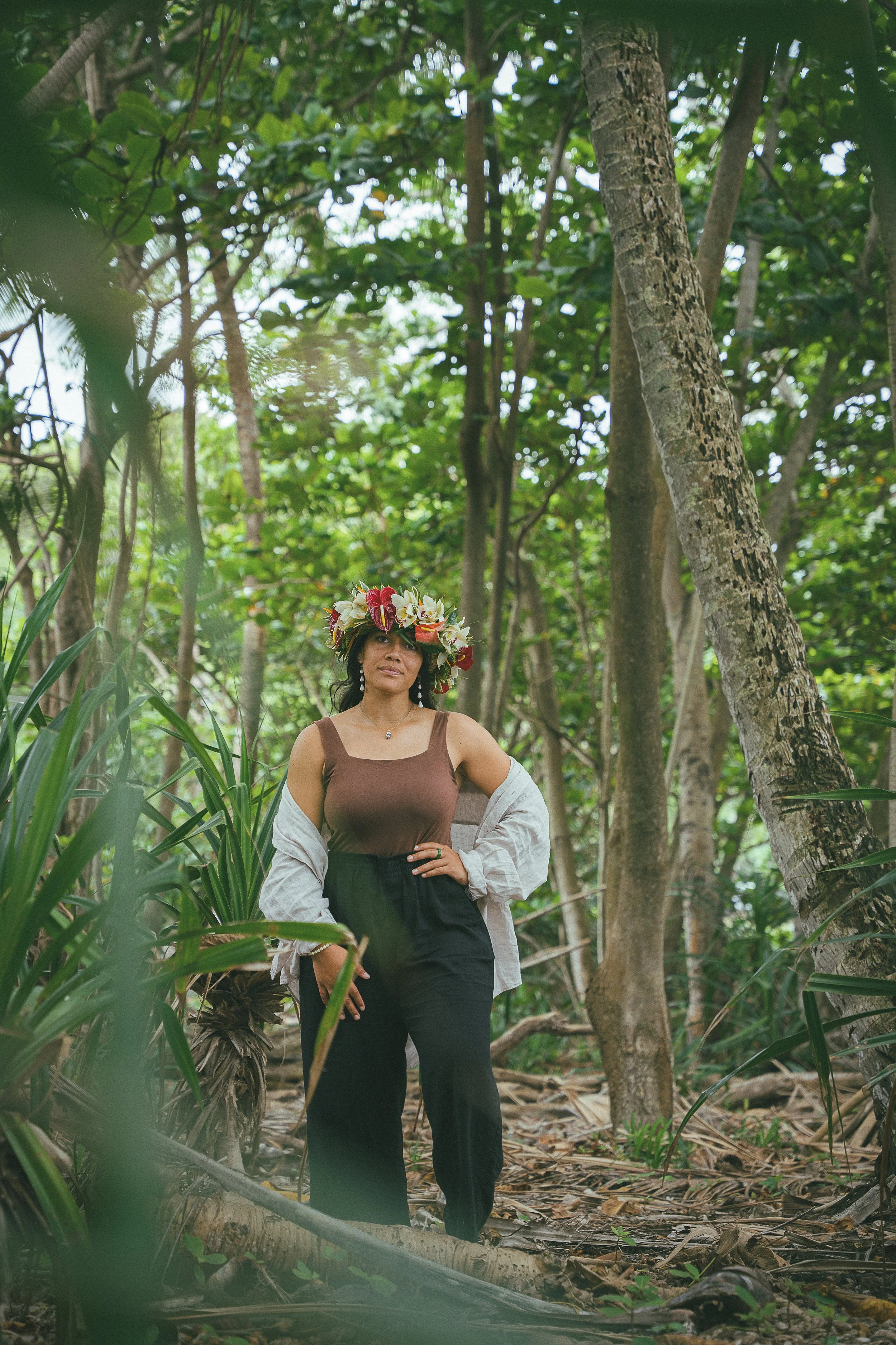 A woman in a brown top, black pants, and a white jacket draped over her shoulders stands in a lush green forest with tall trees and dense foliage. She is wearing a flower crown and jewelry, and has a serene expression on her face.