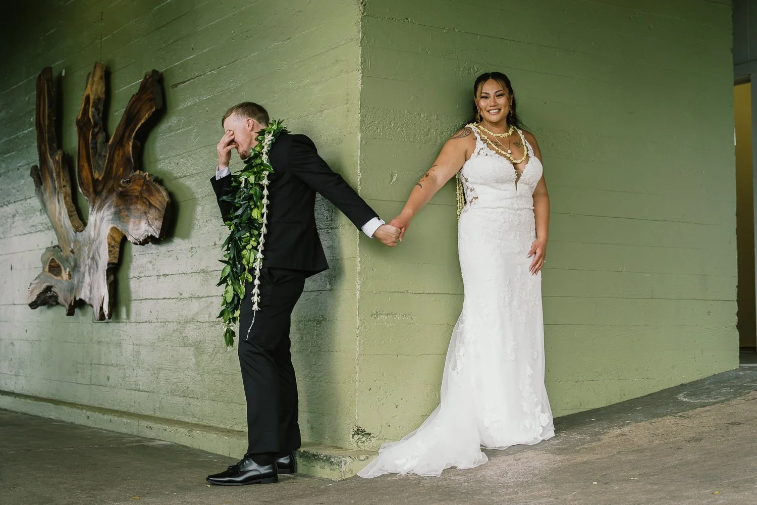 Bride and groom holding hands behind a corner with a green wall, the groom covers his face and wears a lei, the bride smiles in a white lace dress with jewelry.