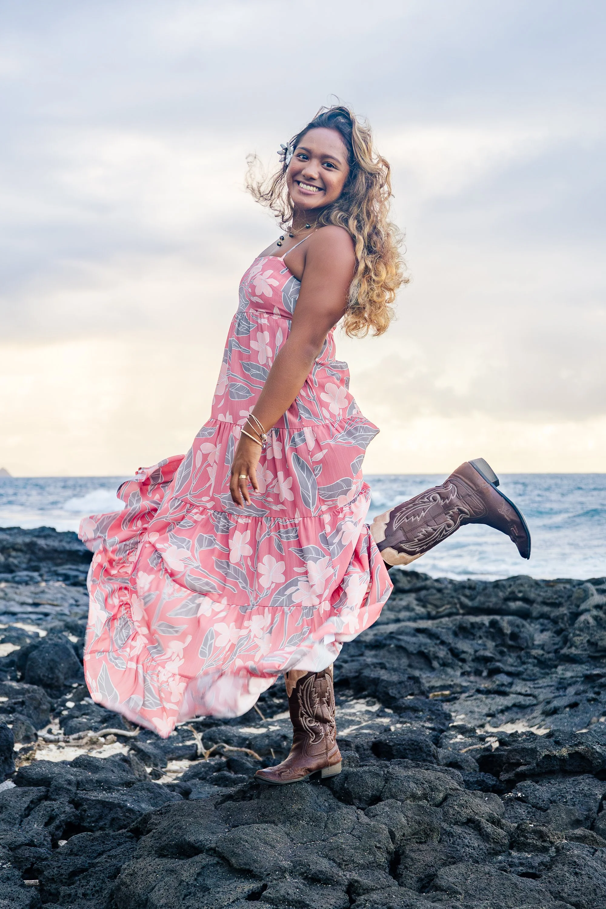 A woman in a pink floral dress and cowboy boots smiling and playing on black volcanic rocks by the ocean at sunset.