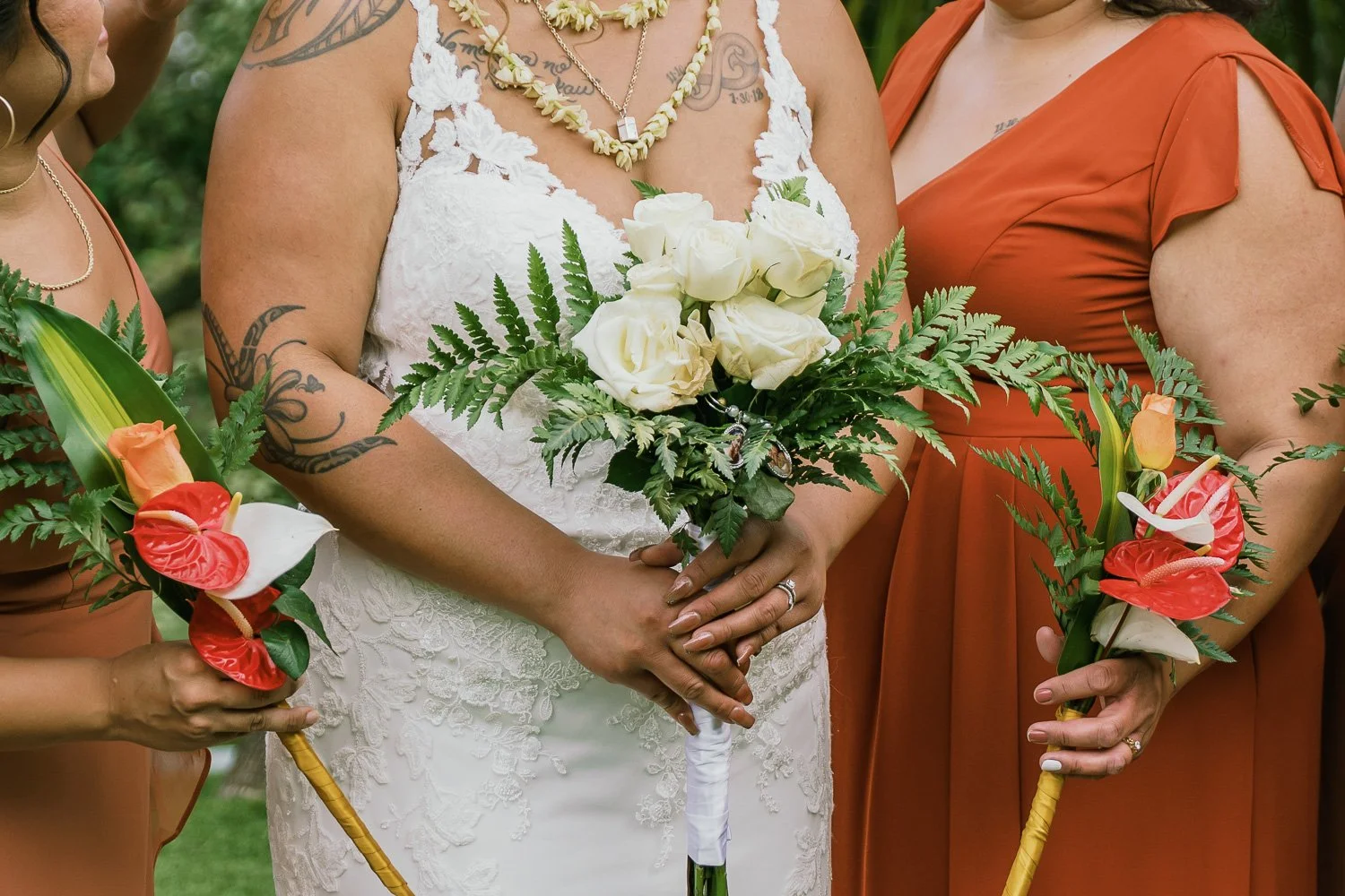 Close-up of a bride in a white lace wedding dress holding a bouquet of white roses and greenery, flanked by bridesmaids in rust-colored dresses holding floral arrangements with orange, white, and red flowers and greenery.