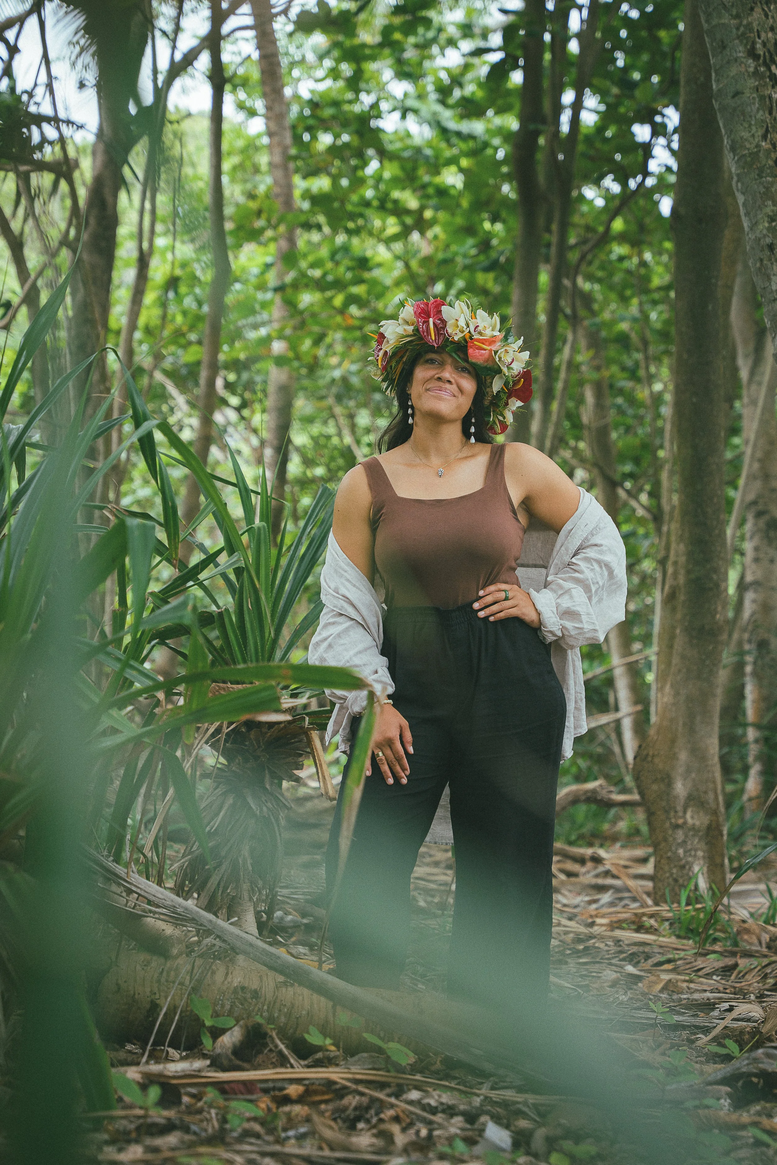 A woman wearing a flower crown, brown tank top, and black pants, standing in a lush green forest with trees and plants around her, with a serene expression on her face.