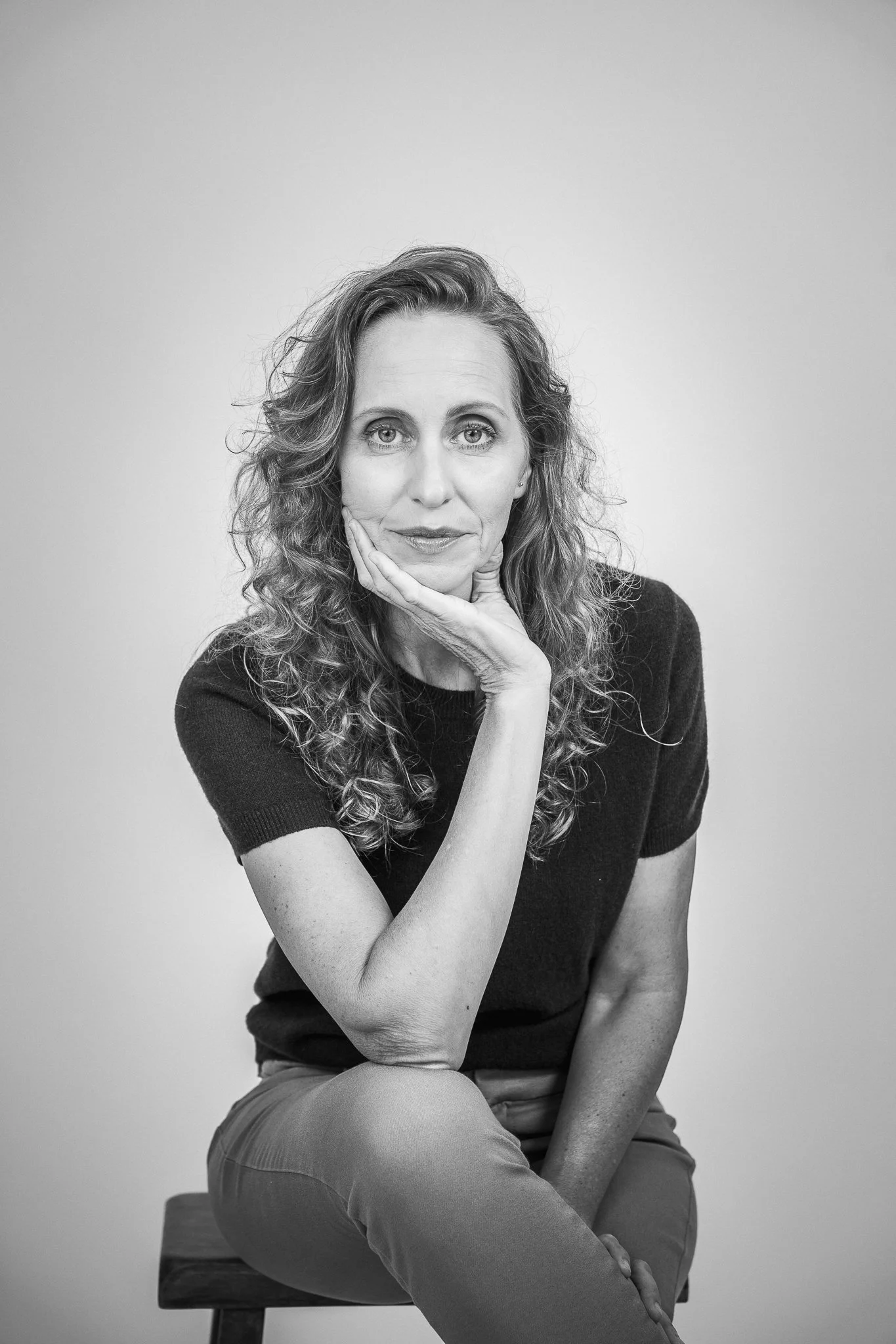 Black and white portrait of a middle-aged woman with curly hair, sitting on a stool against a plain background. She has her chin resting on her hand and is looking at the camera.