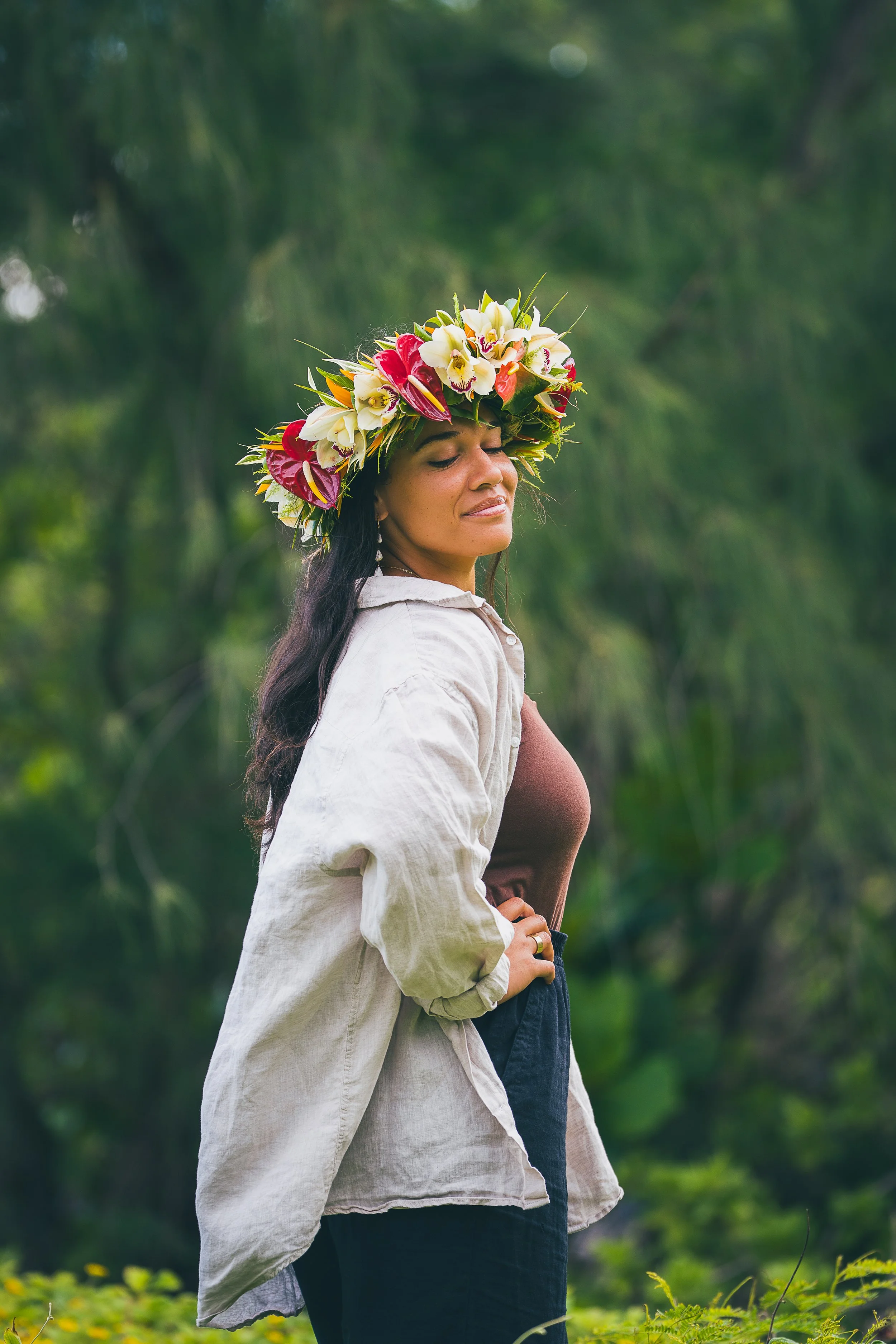 A woman standing outdoors in a natural setting with trees and greenery in the background, wearing a floral crown made of white, pink, and red flowers, a white jacket, and a brown top.