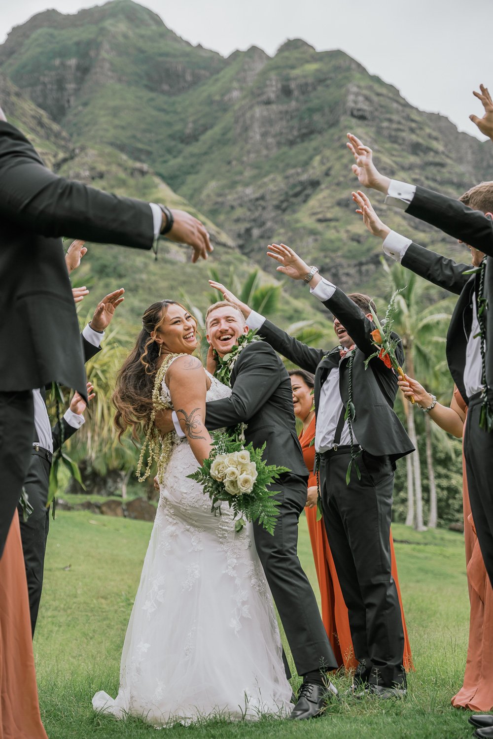 A bride and groom smiling and embracing each other as they are surrounded by friends at a wedding ceremony outdoors, with lush green mountains in the background.