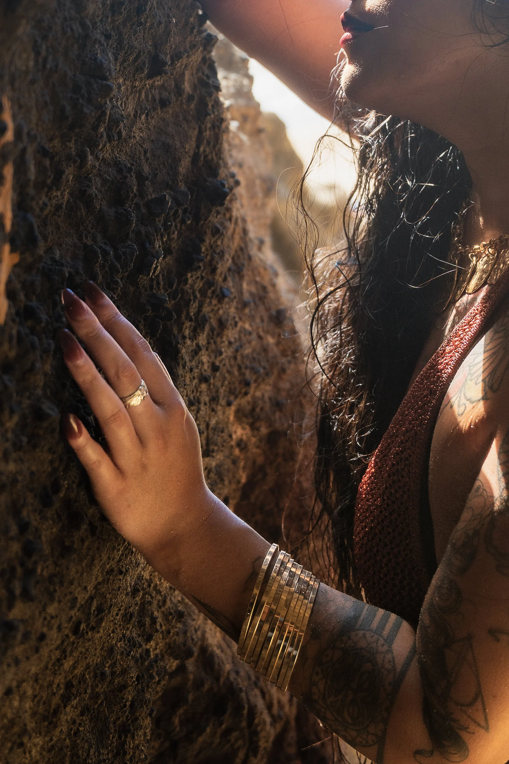 A woman with tattoos and jewelry touching a textured rock wall in the sunlight.