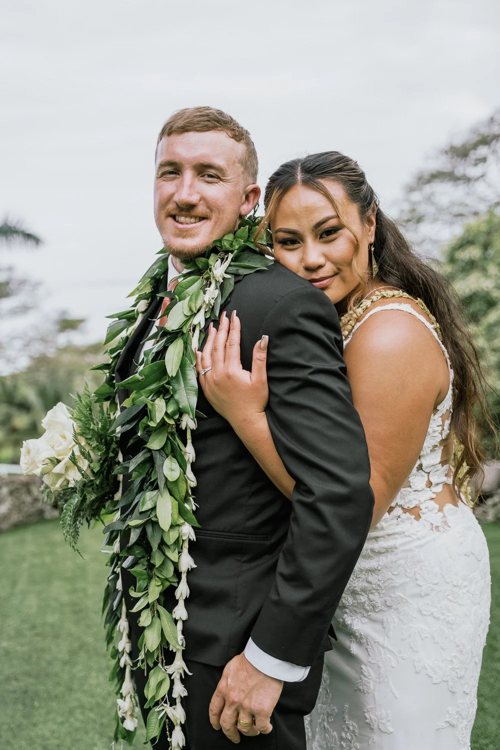 A newlywed couple on their wedding day, with the bride hugging the groom from behind. The groom is wearing a black suit with a leafy garland around his neck, and the bride is in a white lace wedding dress, embracing him affectionately outdoors.