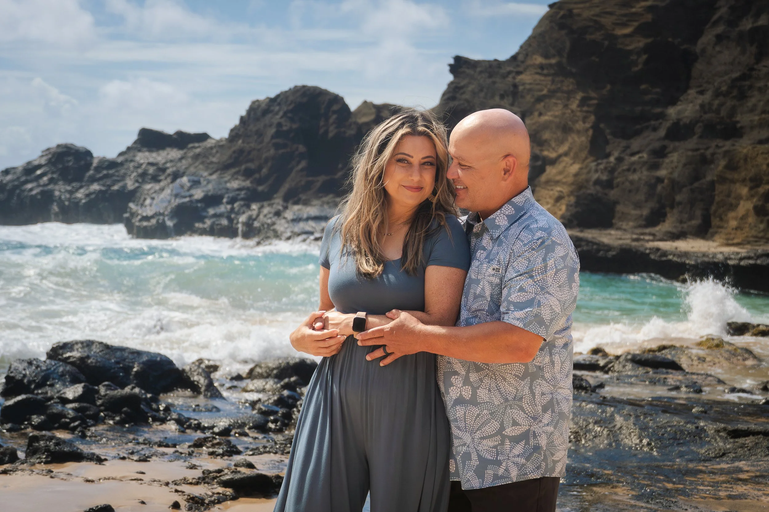 A couple embracing on a rocky beach with ocean waves and cliffs in the background.