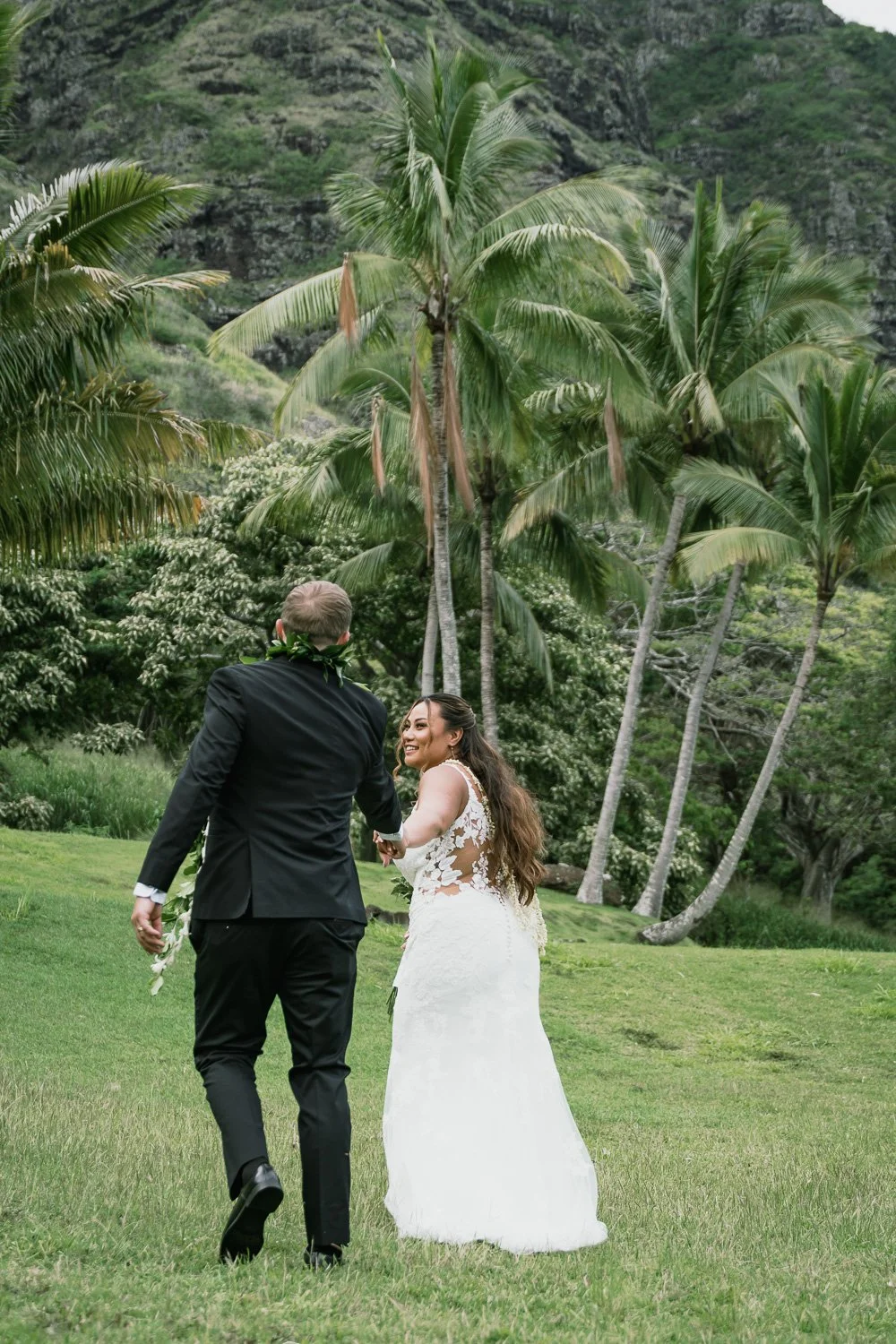 A bride and groom holding hands and smiling in a lush green tropical area with palm trees and mountains in the background.